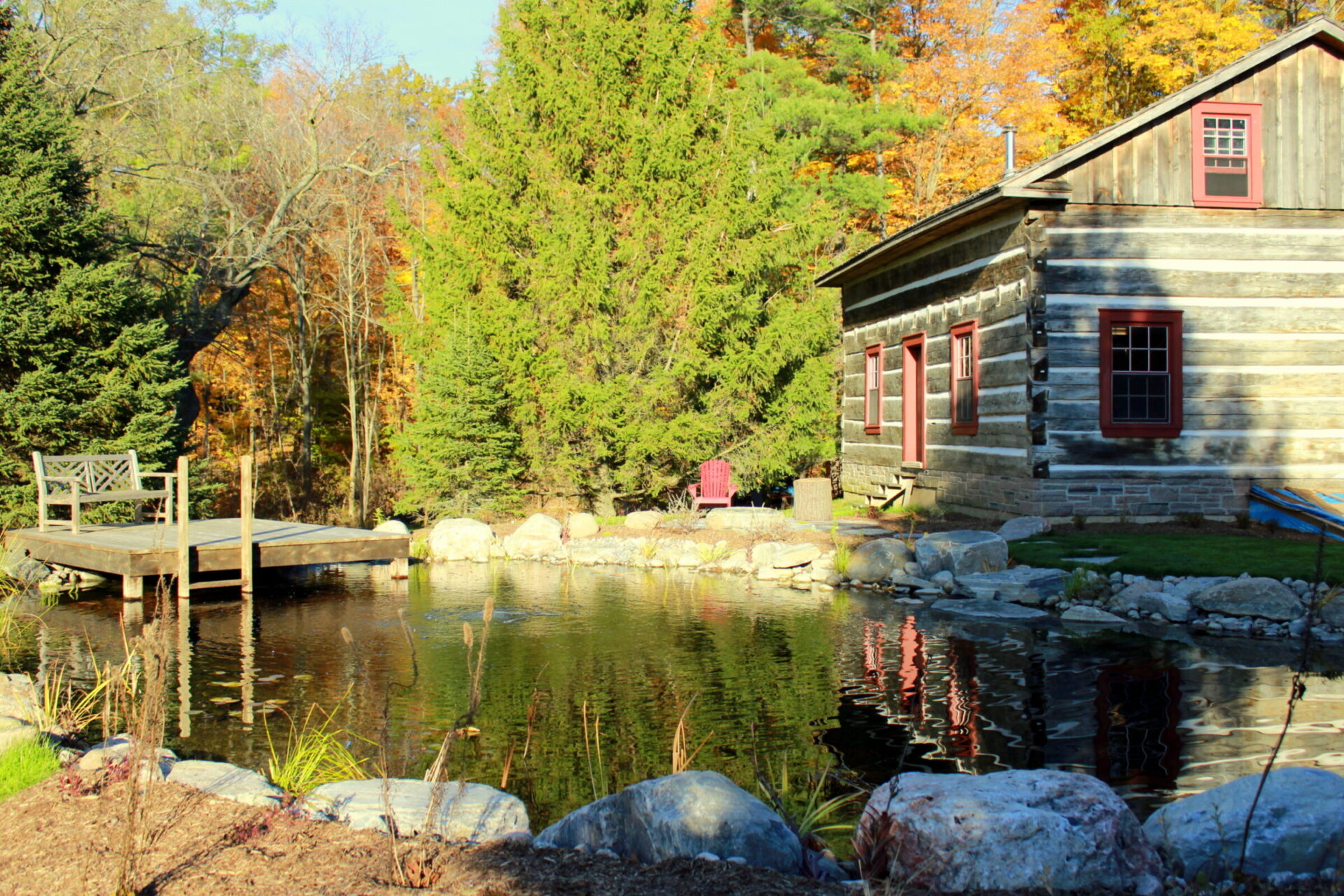 A serene autumn scene with a rustic cabin next to a tranquil pond, surrounded by trees with fall colors, a wooden dock, and a red chair.