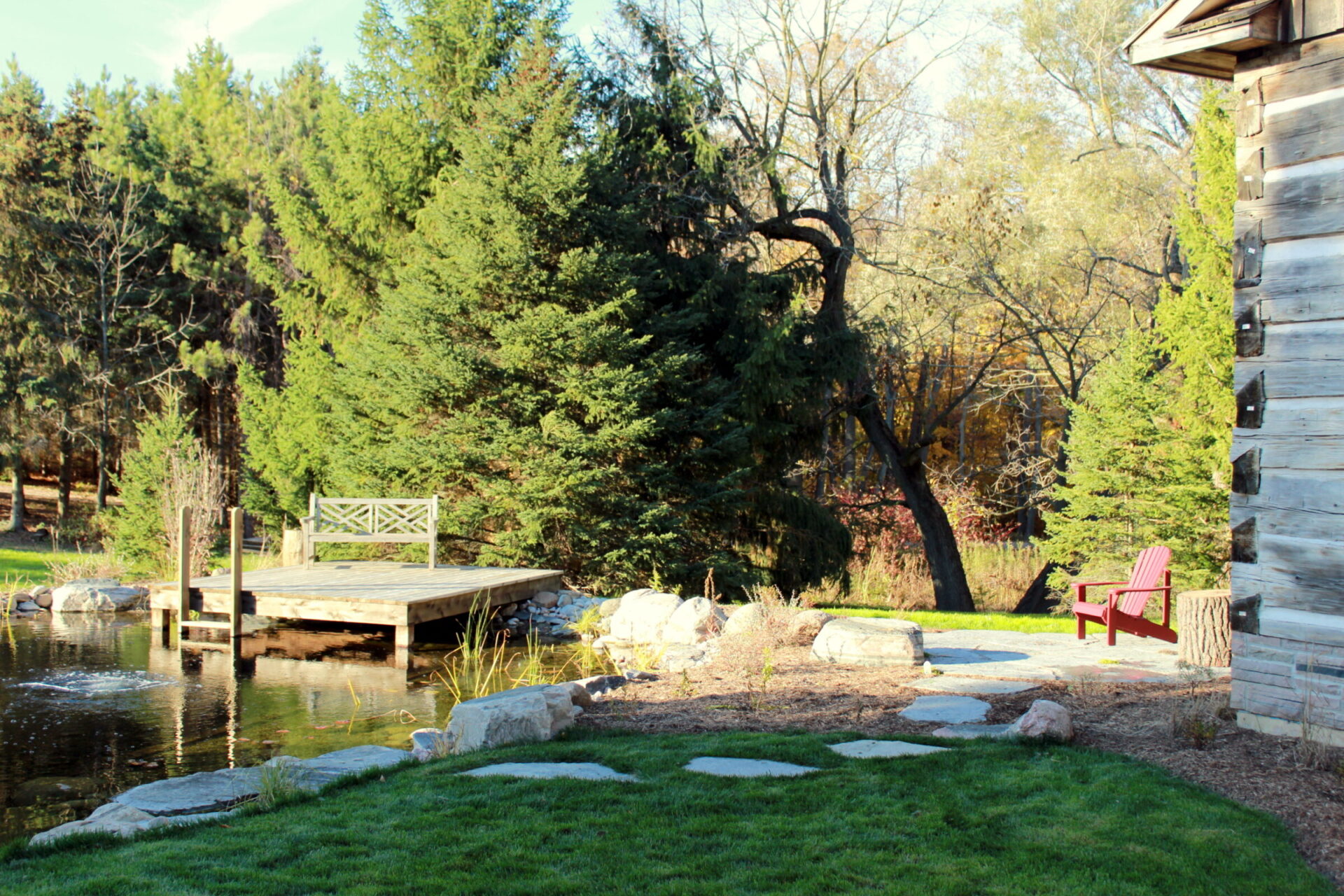 A serene backyard with a wooden dock on a pond, red Adirondack chair, stepping stones, lush greenery, and a rustic cabin side.