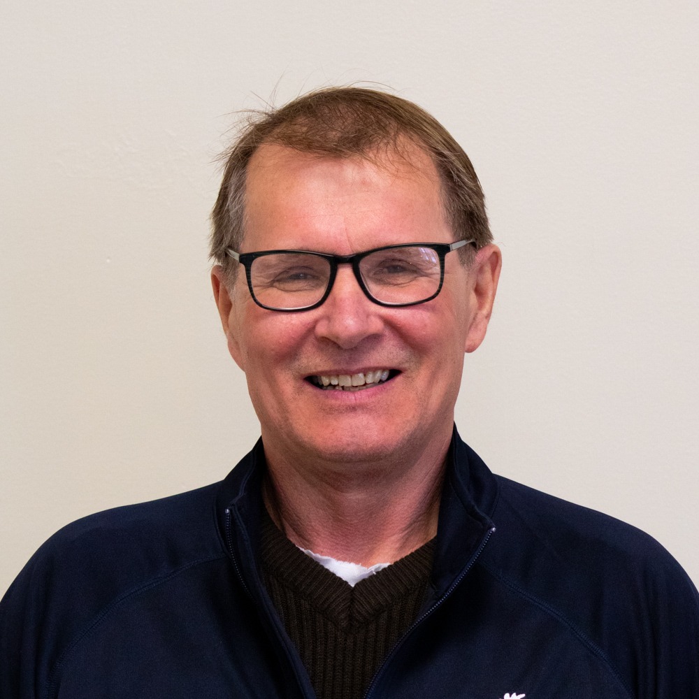 A person is smiling at the camera, wearing glasses, a dark zip-up jacket, and a brown sweater, with a plain light background.