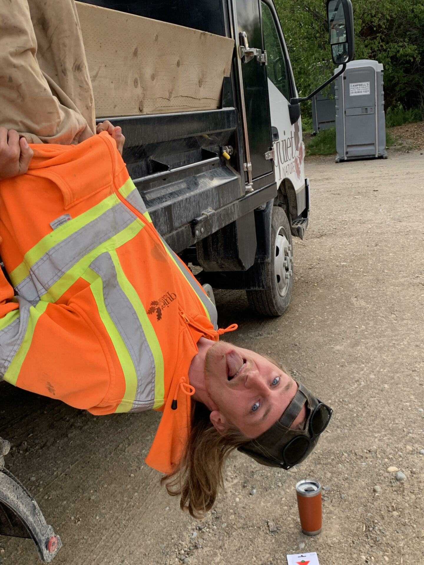 A person wearing a high-visibility jacket is playfully hanging upside down, held by another person, with a construction truck and portable toilet in the background.