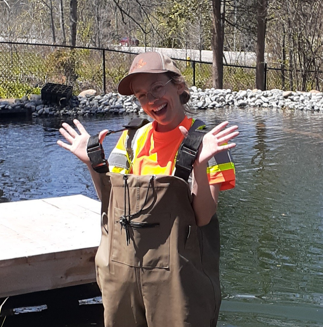 A person in a high-visibility vest and chest waders is standing on a dock, smiling and gesturing with their hands up, near water.