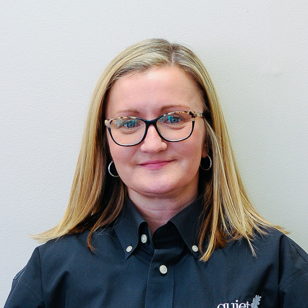 The image displays a smiling person with blonde hair wearing glasses, hoop earrings, and a dark buttoned shirt against a light background.