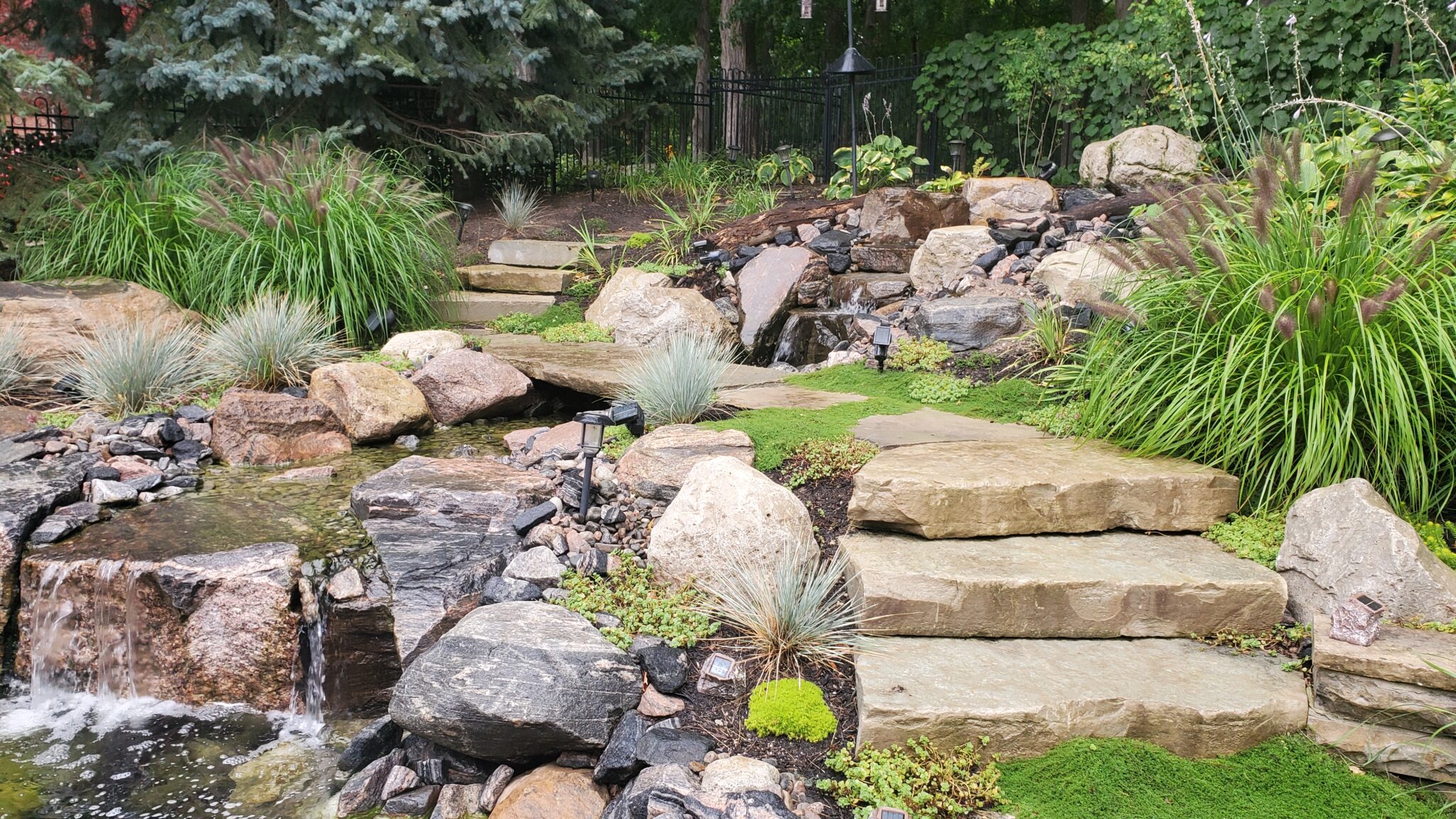 This image displays a landscaped garden featuring a small waterfall, stone steps, lush greenery, ornamental grasses, and a variety of rocks, creating a serene outdoor space.