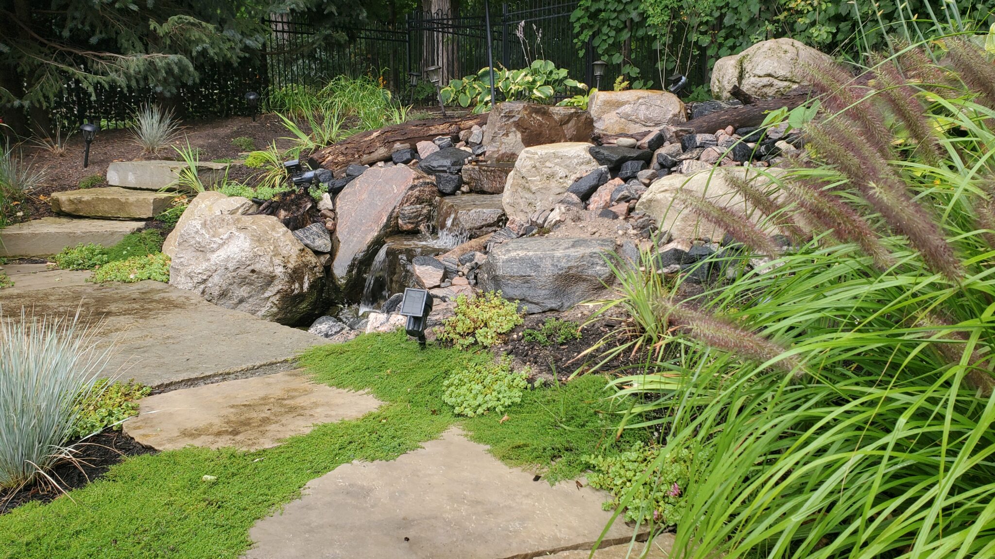 This image shows a landscaped garden with a rock waterfall, lush greenery, a path, and ornamental grasses. It appears tranquil and well-maintained.