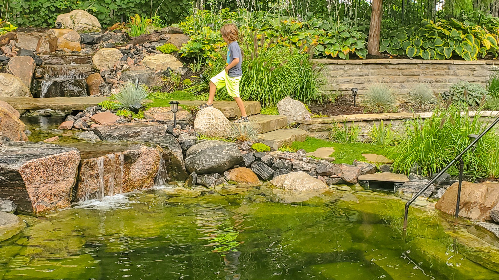 A child walks on rocks beside a small garden waterfall and pond. Lush green vegetation surrounds the tranquil water feature. Outdoor lighting accents the scene.