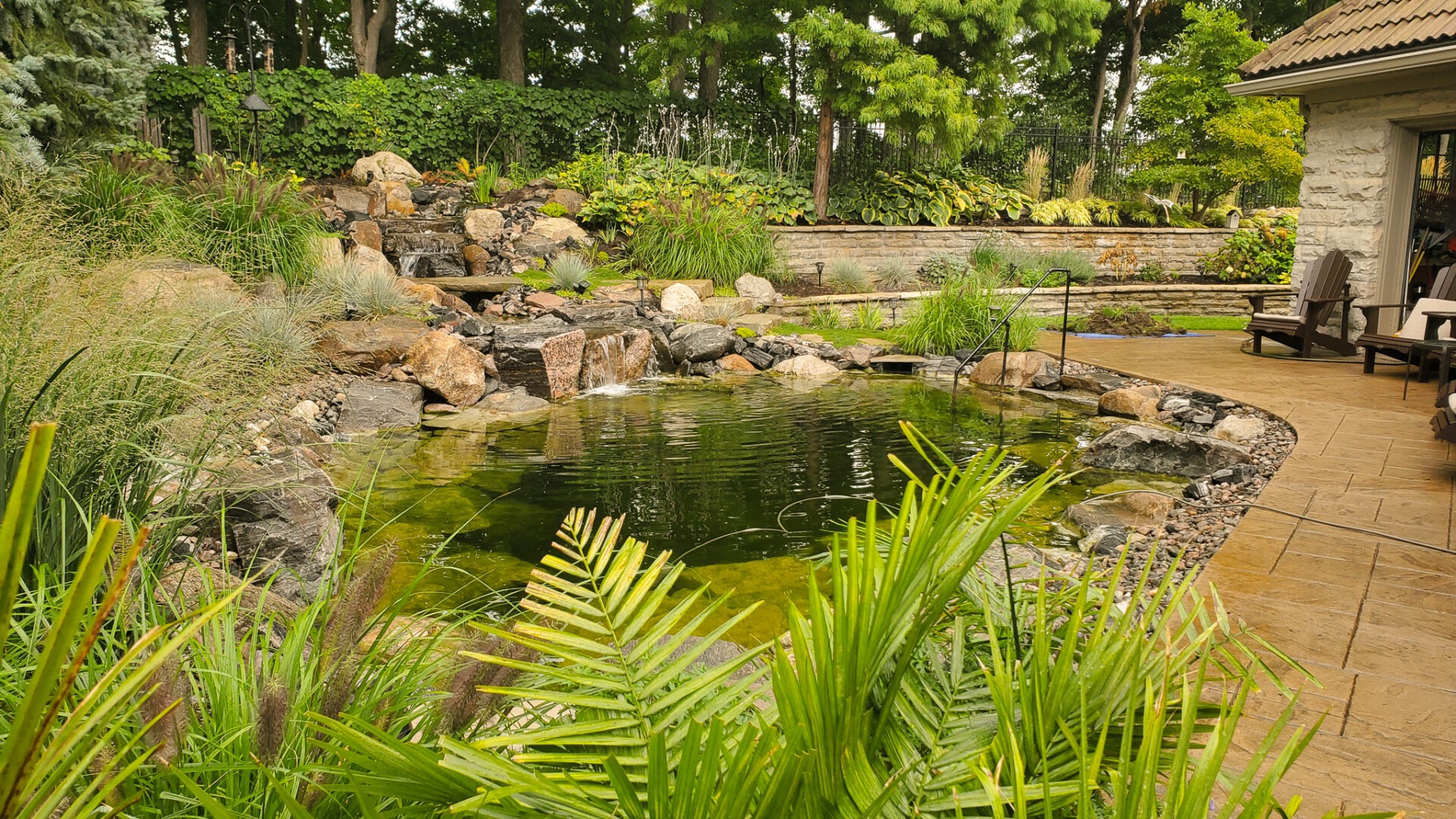 A serene backyard garden with a pond, surrounded by lush plants, rocks, a small waterfall, and a stone patio area with seating.