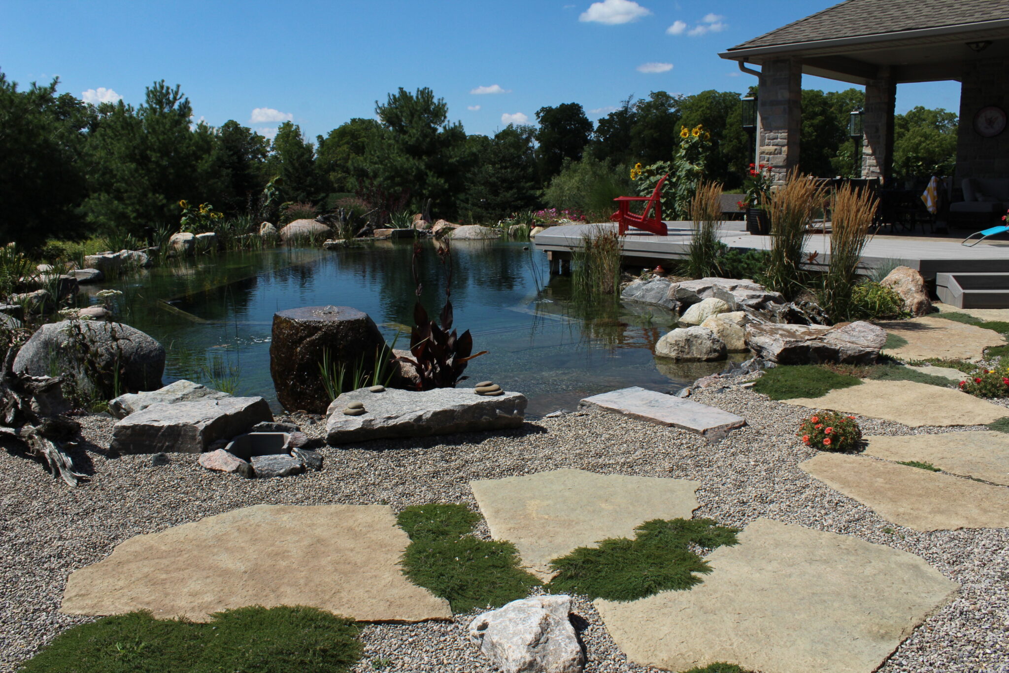 This image shows a landscaped backyard with a naturalistic pond, stone pathways, boulders, greenery, and a gazebo with a red chair, under clear skies.