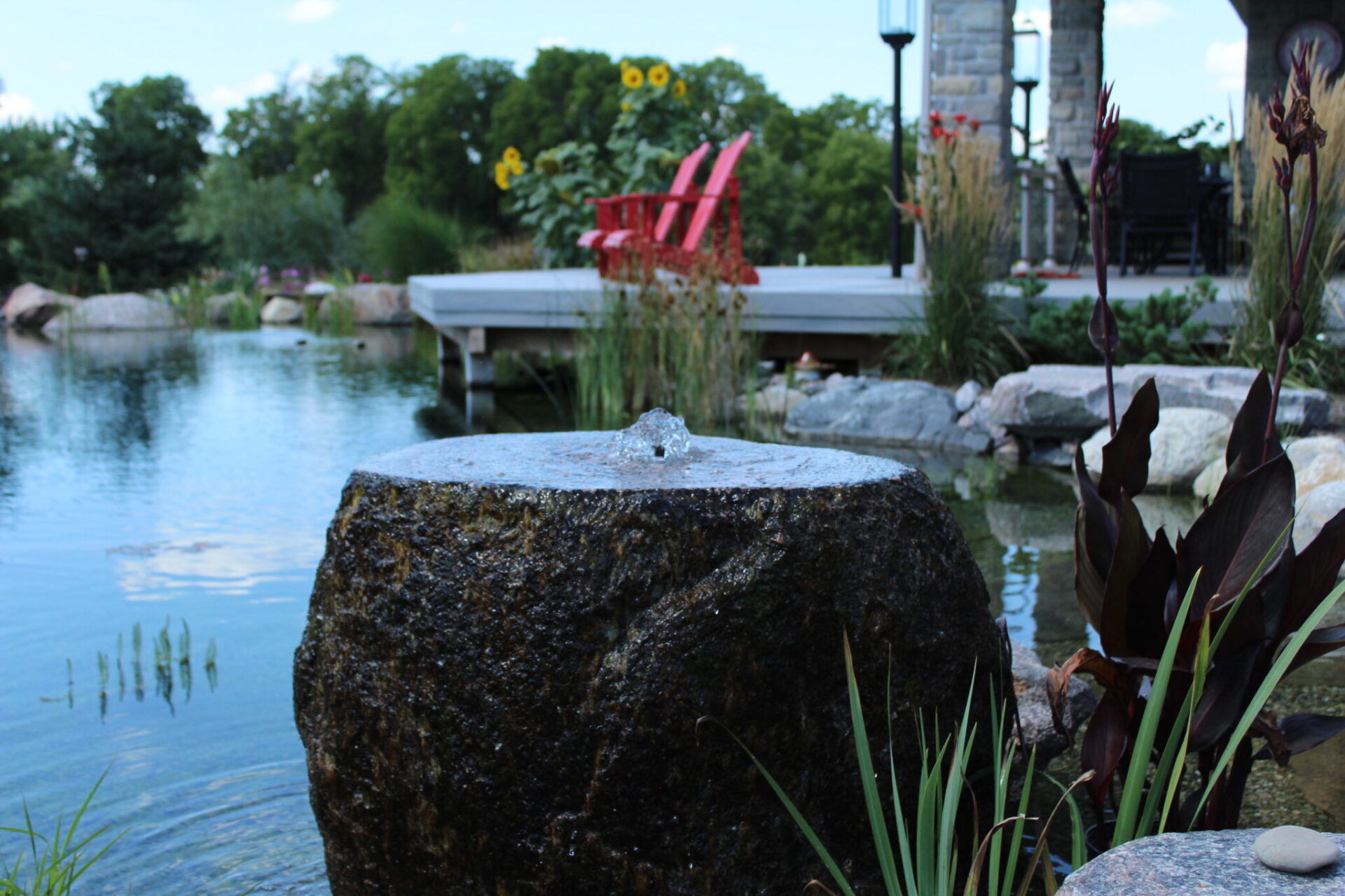 A tranquil pond with a bubbling fountain stone in the foreground, surrounded by greenery, with a wooden dock and red chairs in the background.