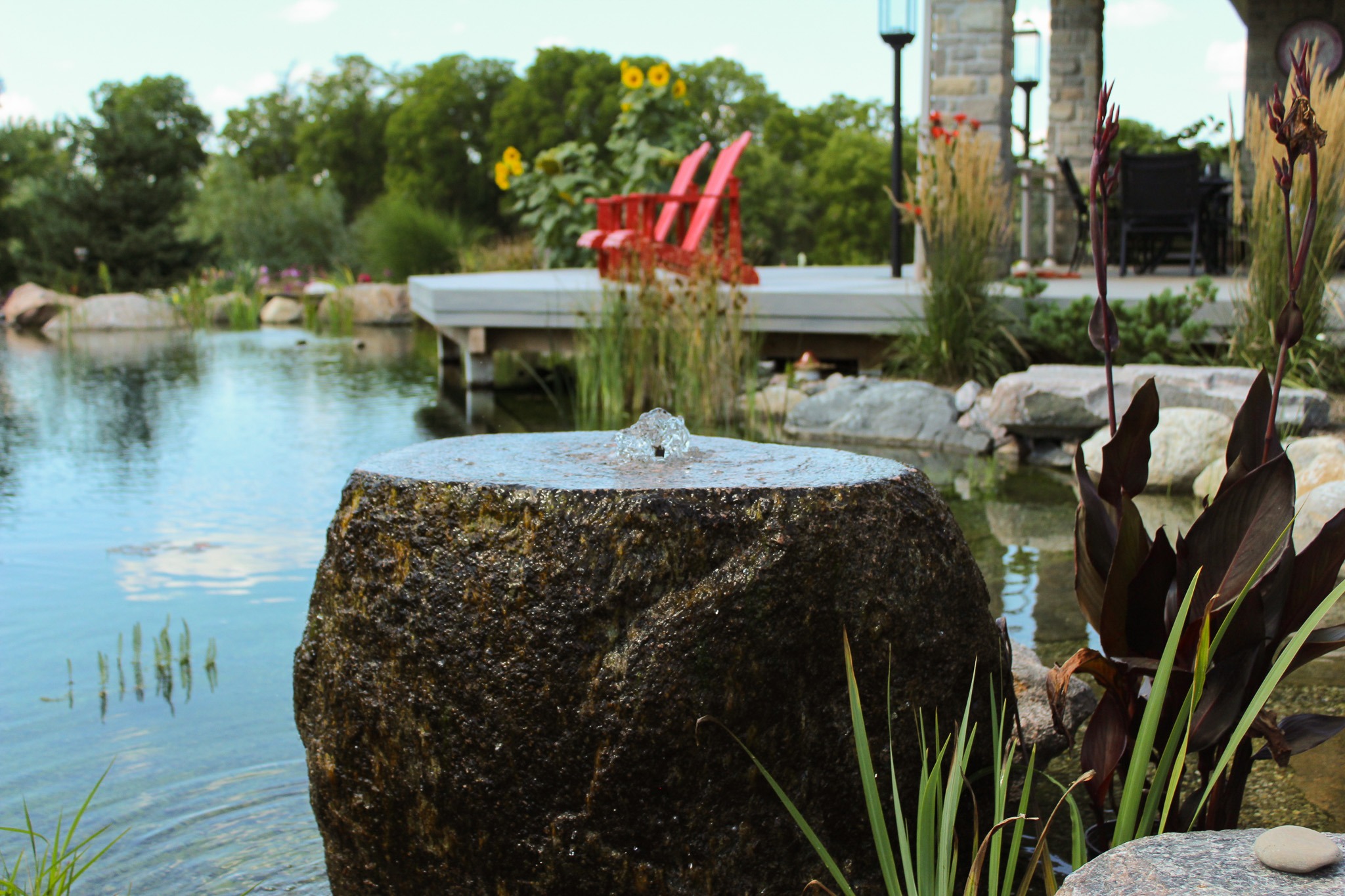 This image features a tranquil garden with a pond, rocks, vegetation, and a fountain jetting water atop a boulder, with red Adirondack chairs in the distance.