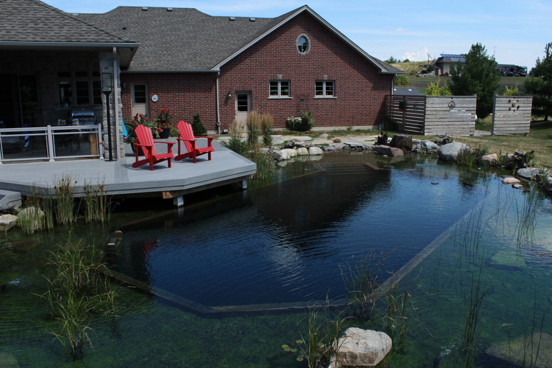 A serene backyard with a brick house, wooden deck, two red chairs, and a clear, landscaped pond surrounded by rocks and plants.