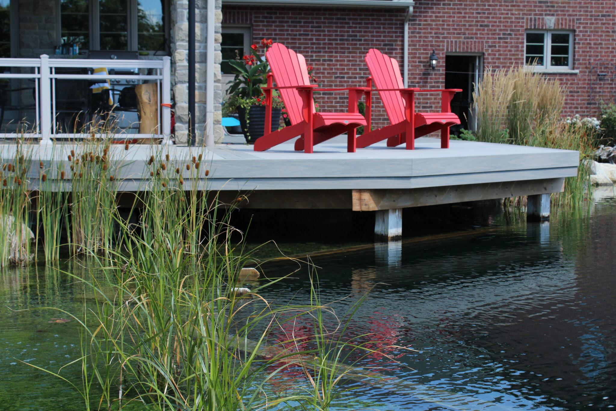 Two red Adirondack chairs are positioned on a wooden dock overlooking a tranquil pond with lush greenery, near a brick building with large windows.