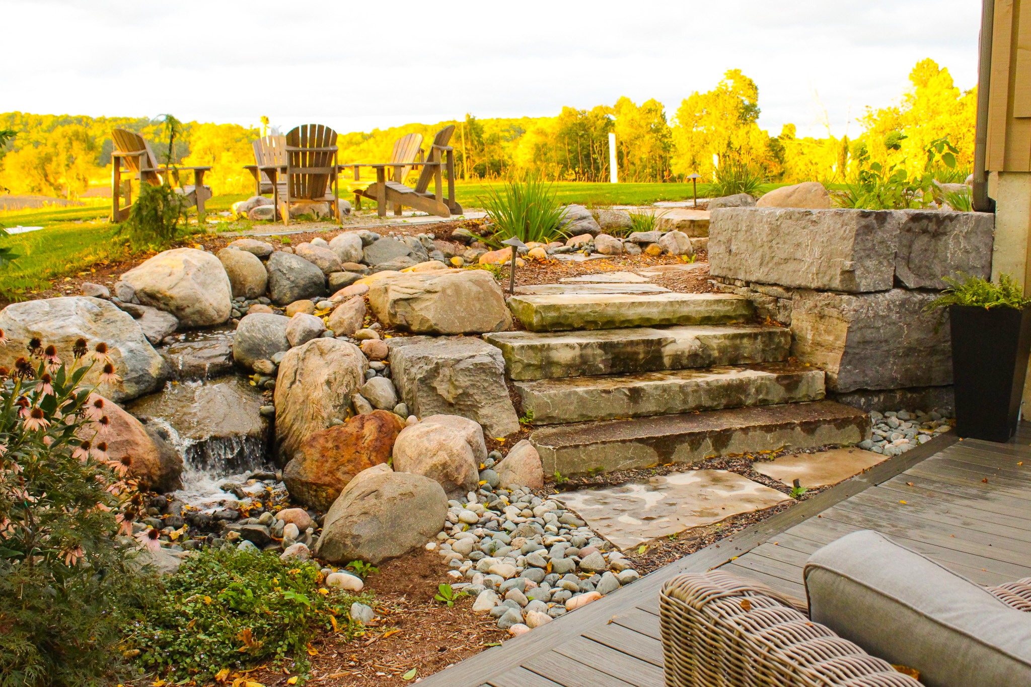 This image shows a tranquil backyard with Adirondack chairs, a stone stairway, a small waterfall, landscaping, and a wooden deck in a serene setting.