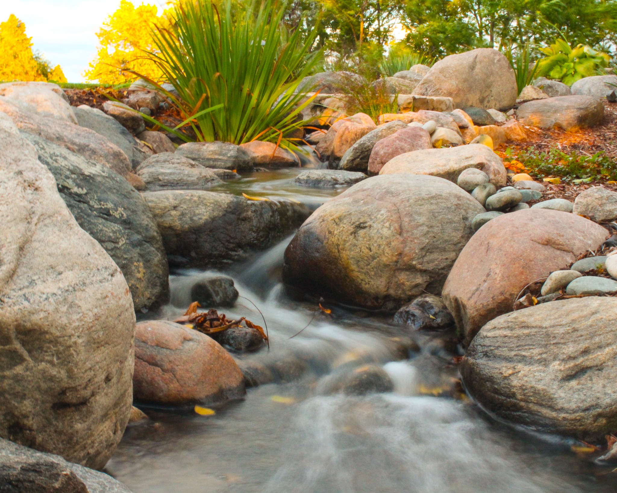 A serene stream flows through large rounded rocks with a soft, mist-like quality, suggesting a long exposure photo; lush greenery and autumn leaves frame the scene.