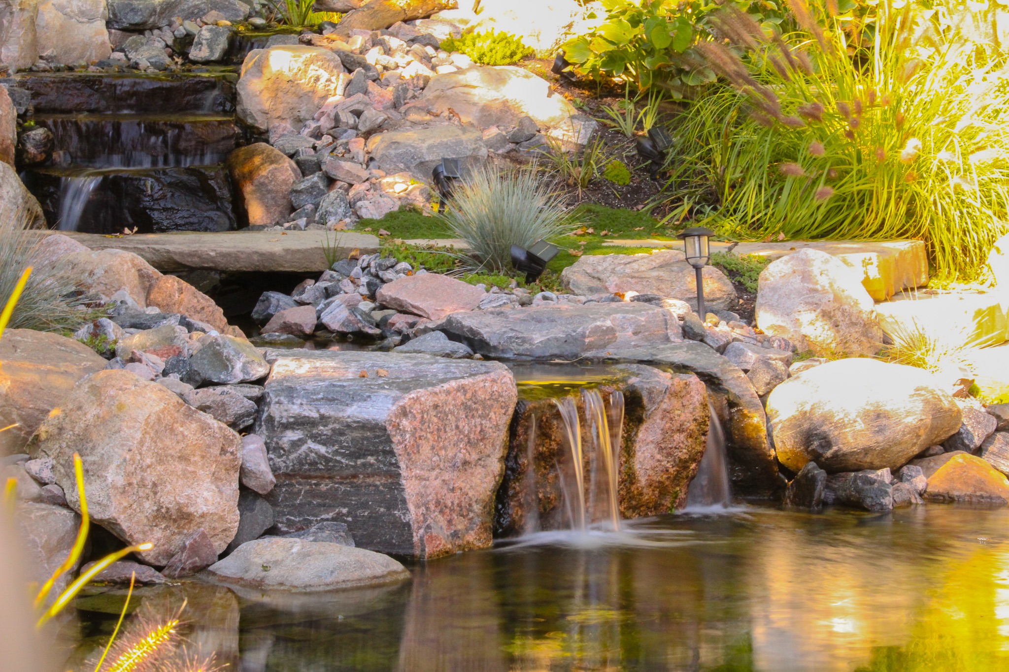 The image shows a serene garden pond with cascading waterfalls over rocks, surrounded by lush greenery and equipped with outdoor lighting features.