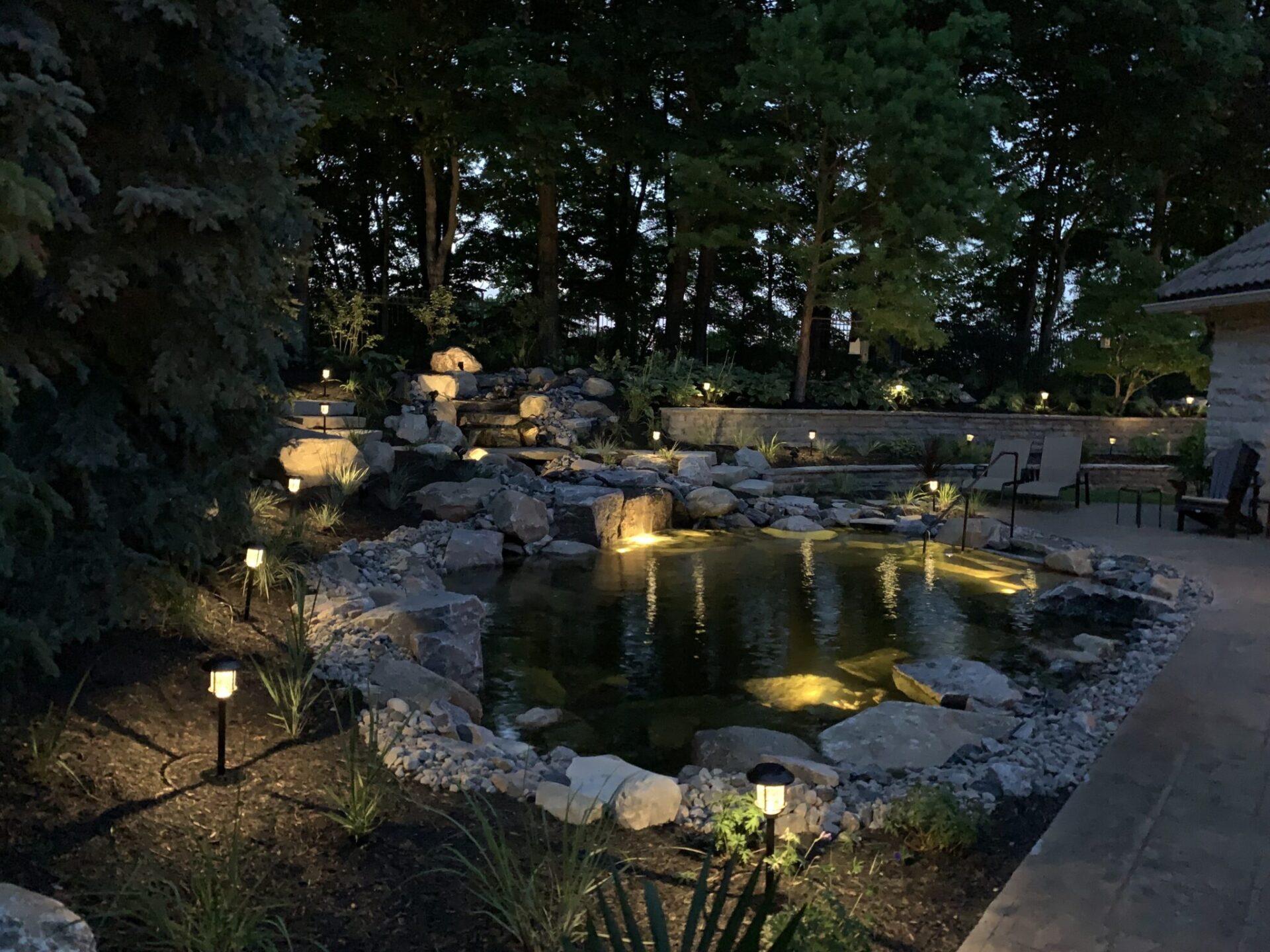 An illuminated garden at dusk with a pond surrounded by rocks and greenery, path lights, benches, and a stone structure to the side.