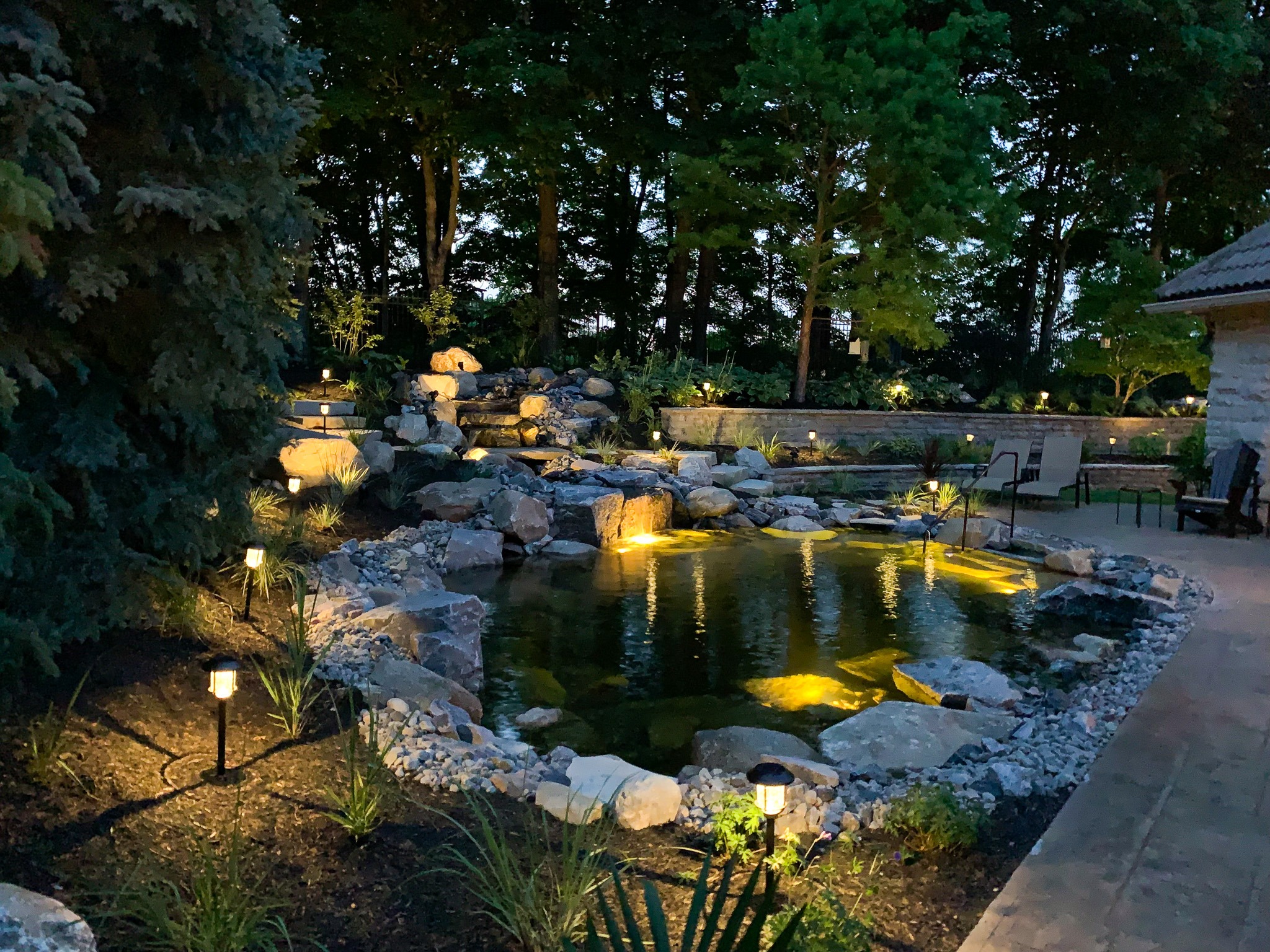 A tranquil backyard garden at dusk with a pond, illuminated by landscape lighting, surrounded by rocks, plants, and a seating area.