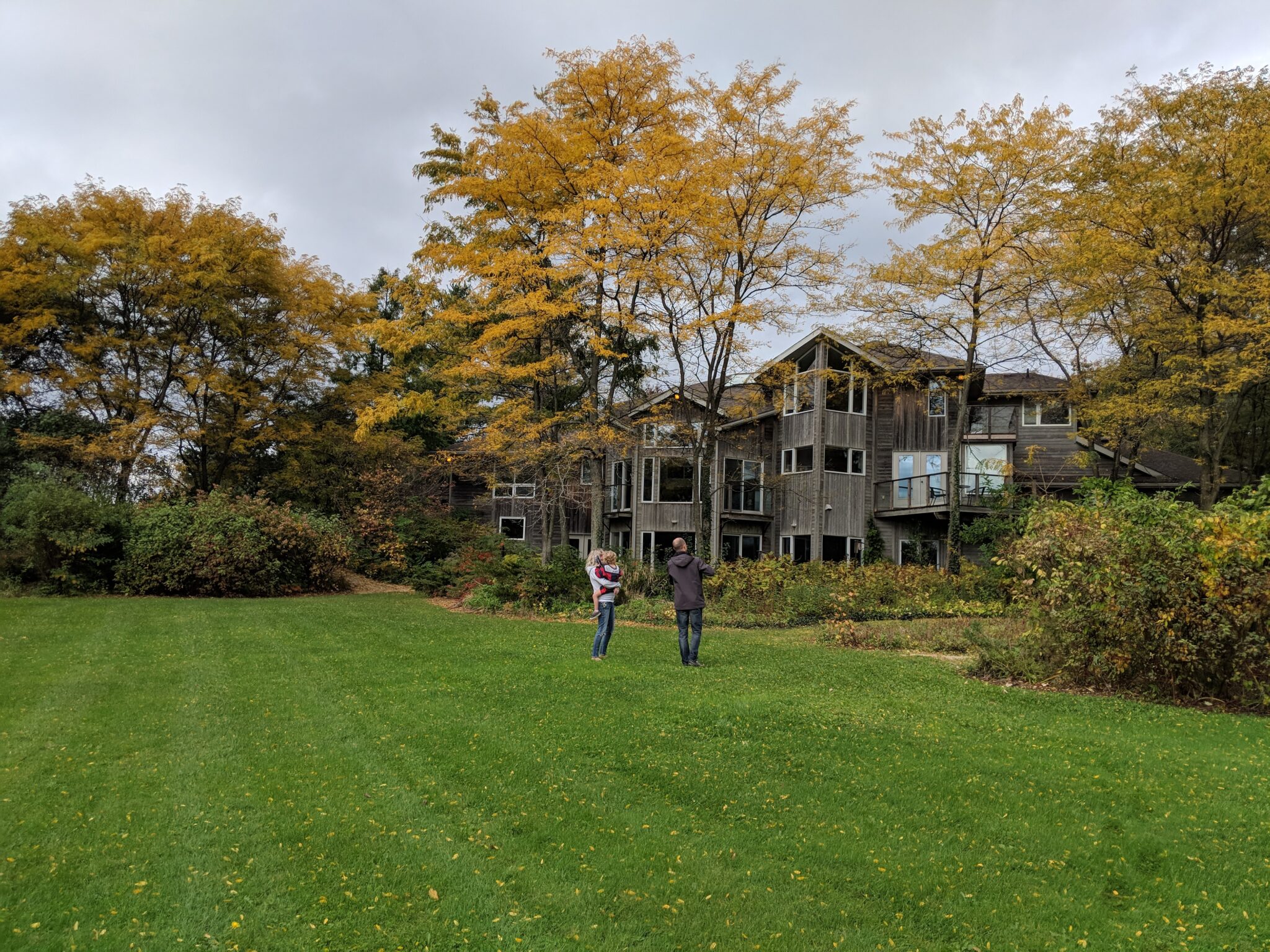 Two people stand on a lush green lawn in front of a large house with wooden features, surrounded by trees with autumn foliage under a cloudy sky.
