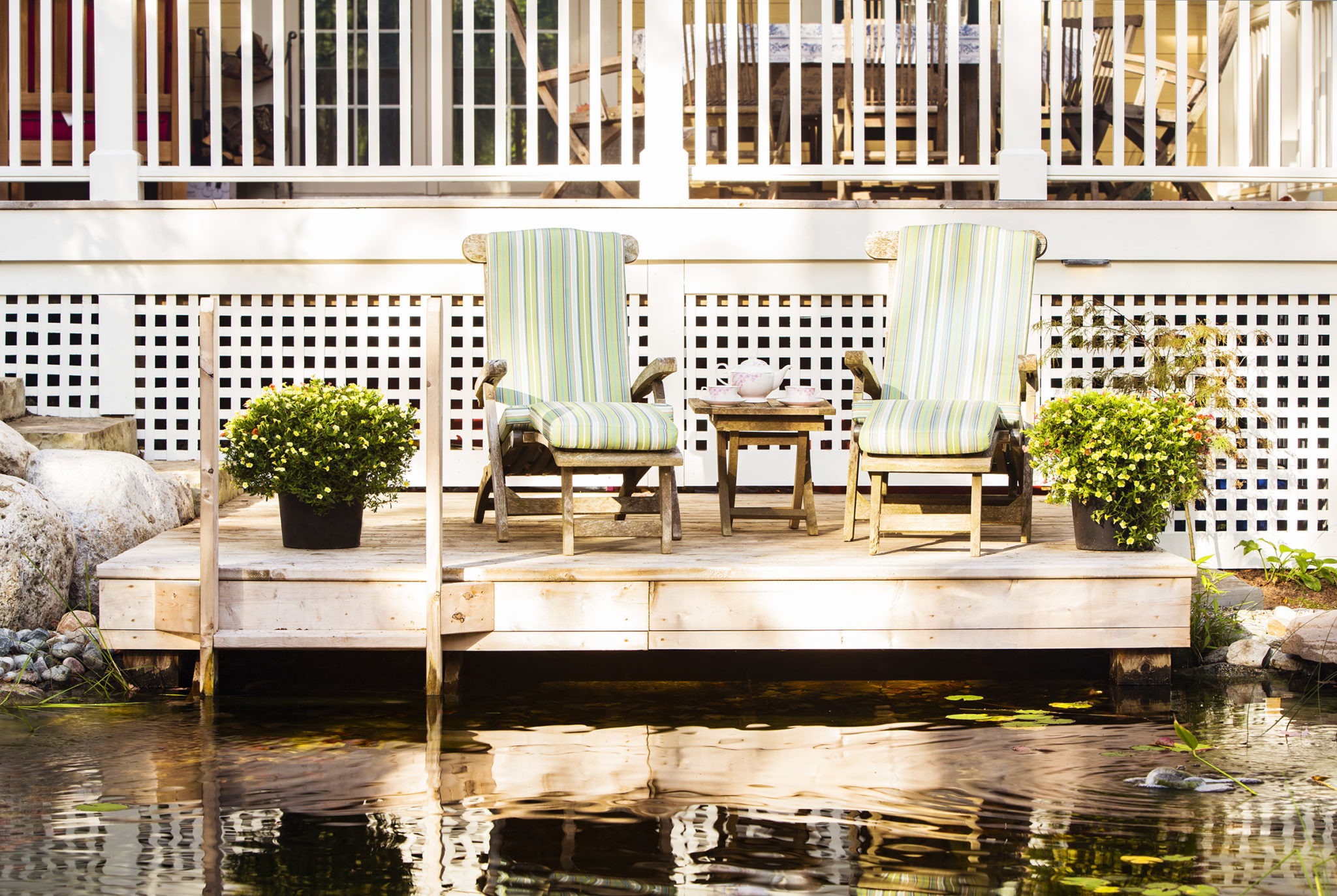 Two striped cushioned armchairs on a wooden deck by a pond, with a small table between them. Plants adorn the tranquil outdoor seating area.