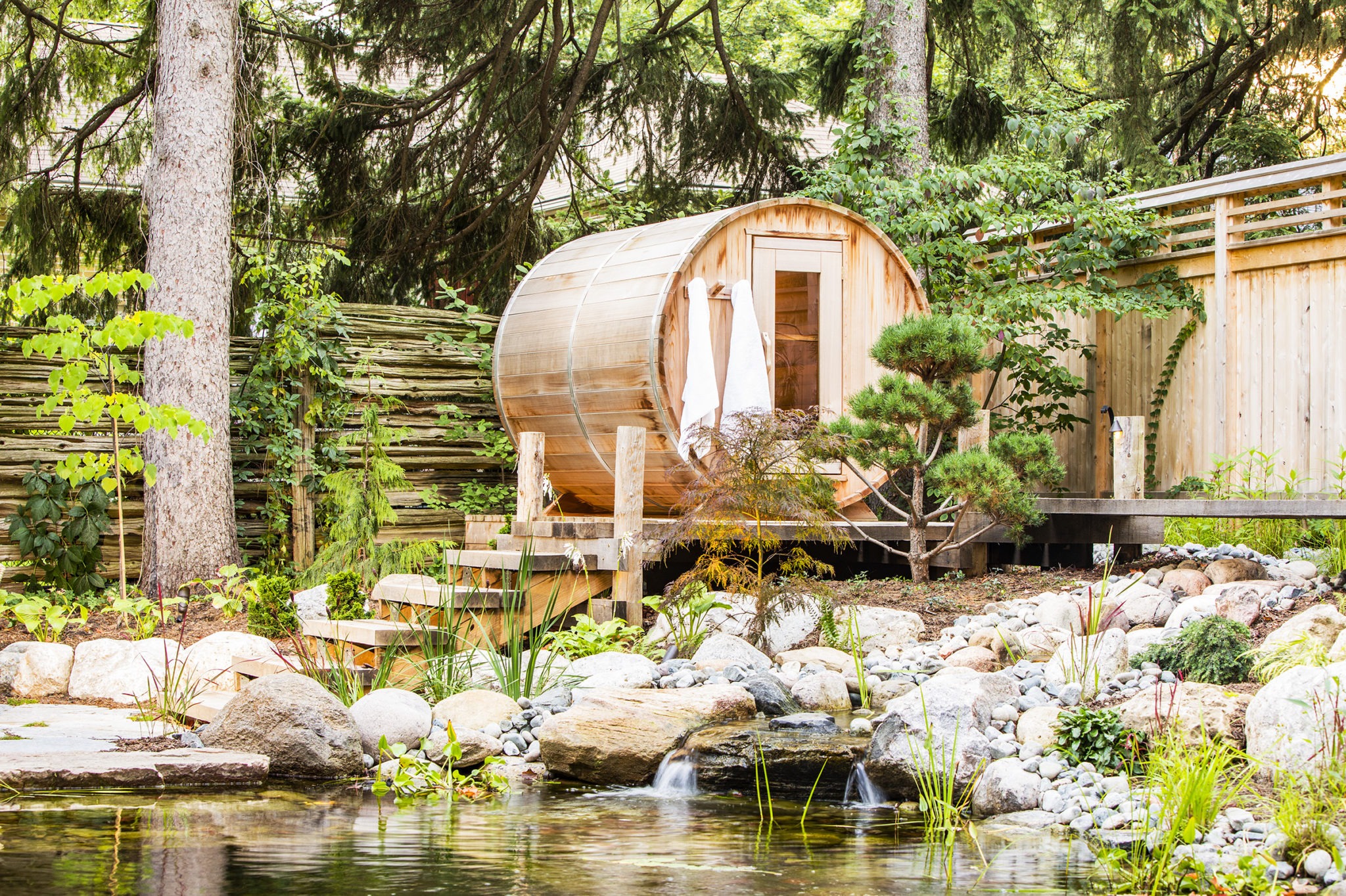 A serene outdoor scene with a wooden barrel sauna by a pond surrounded by lush greenery, rocks, and a small wooden bridge.