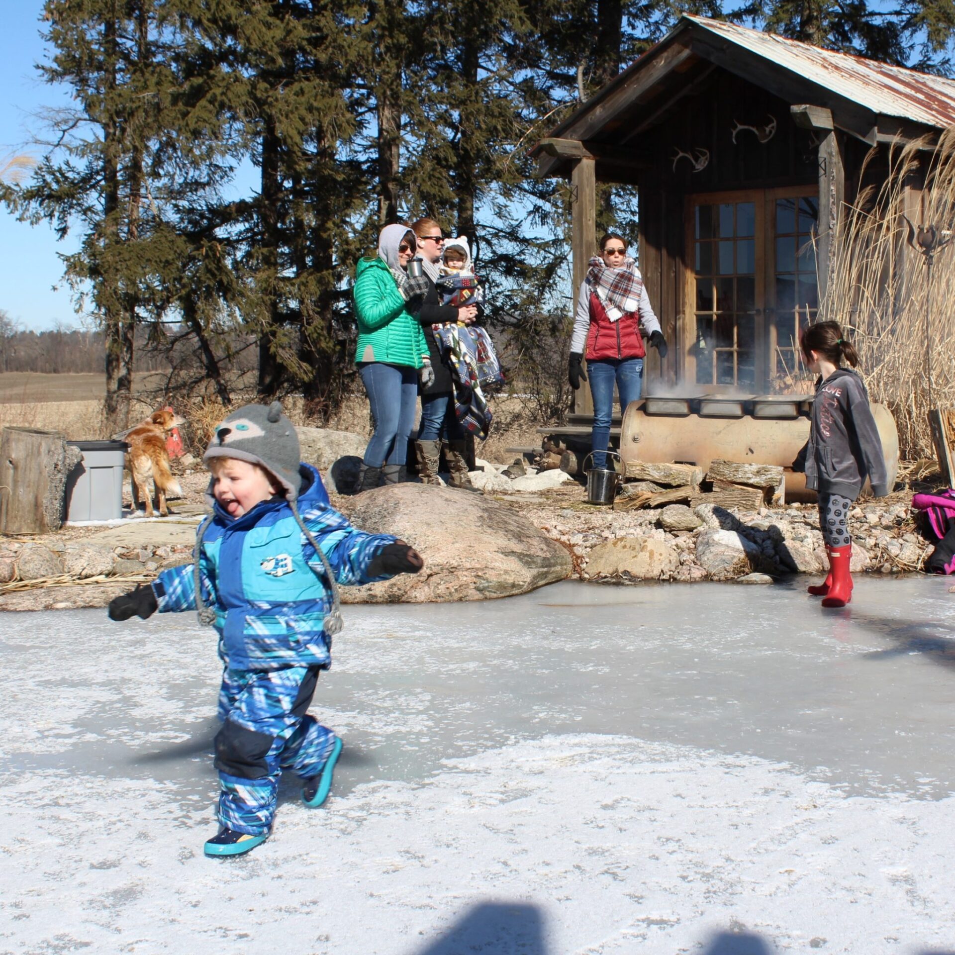 A child in blue winter gear appears delighted, running across a frozen surface outdoors, while several people and a dog stand nearby. A rustic building emits smoke.