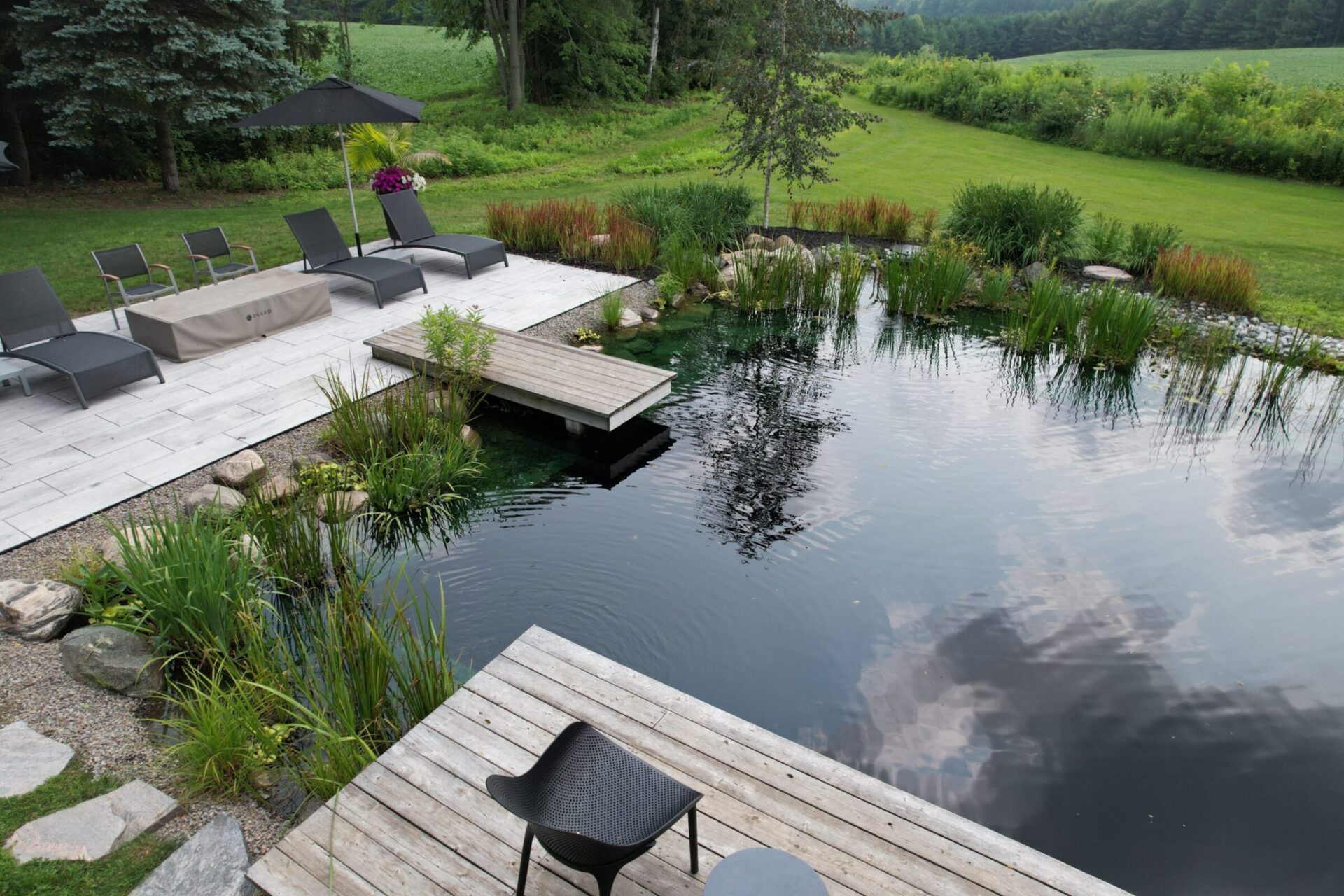 An outdoor relaxation area with sleek lounge chairs, a wooden deck, a serene pond, and lush greenery under a cloudy sky.