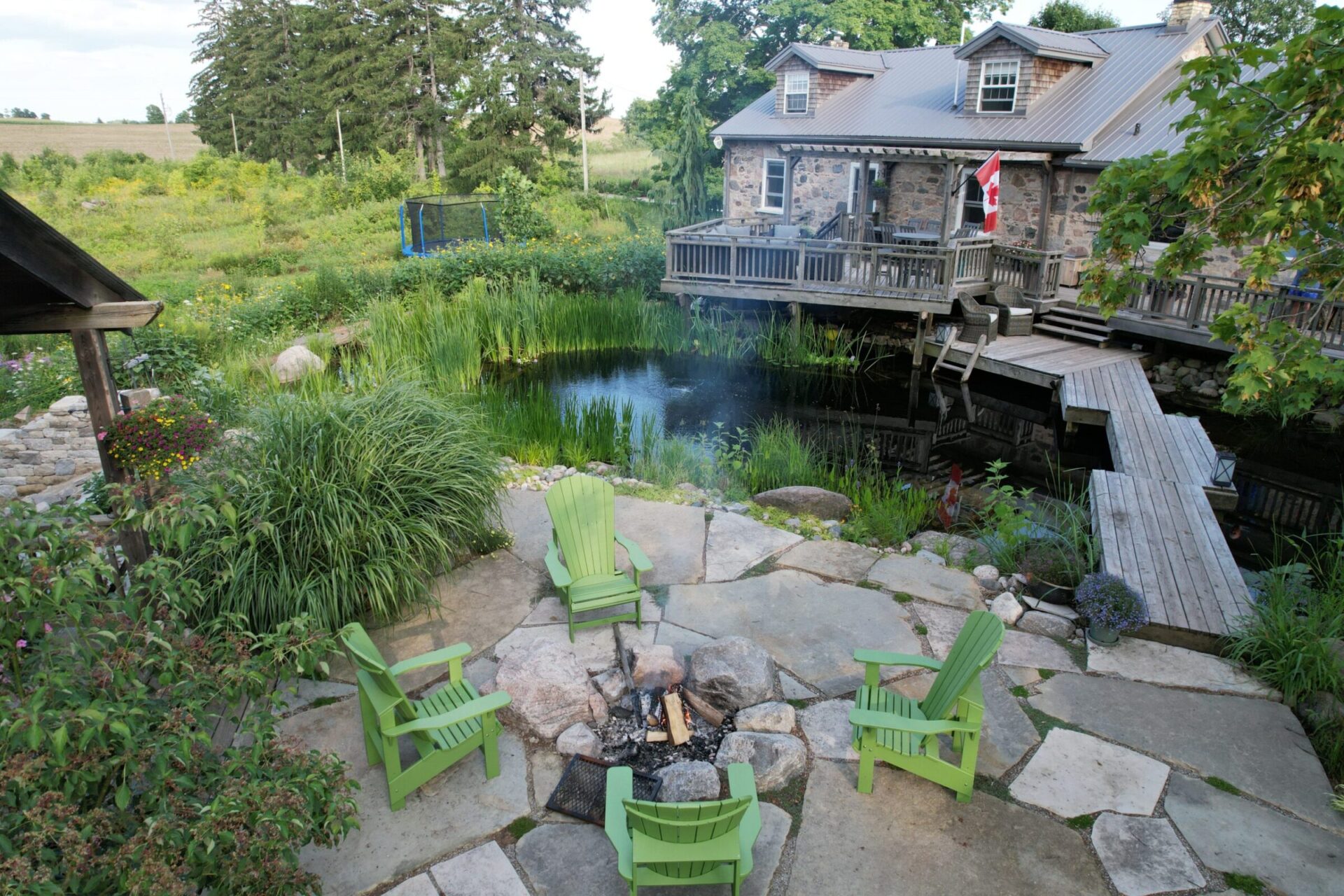 A rustic stone house with a Canadian flag overlooks a tranquil pond, surrounded by lush greenery and a stone patio with fire pit and chairs.