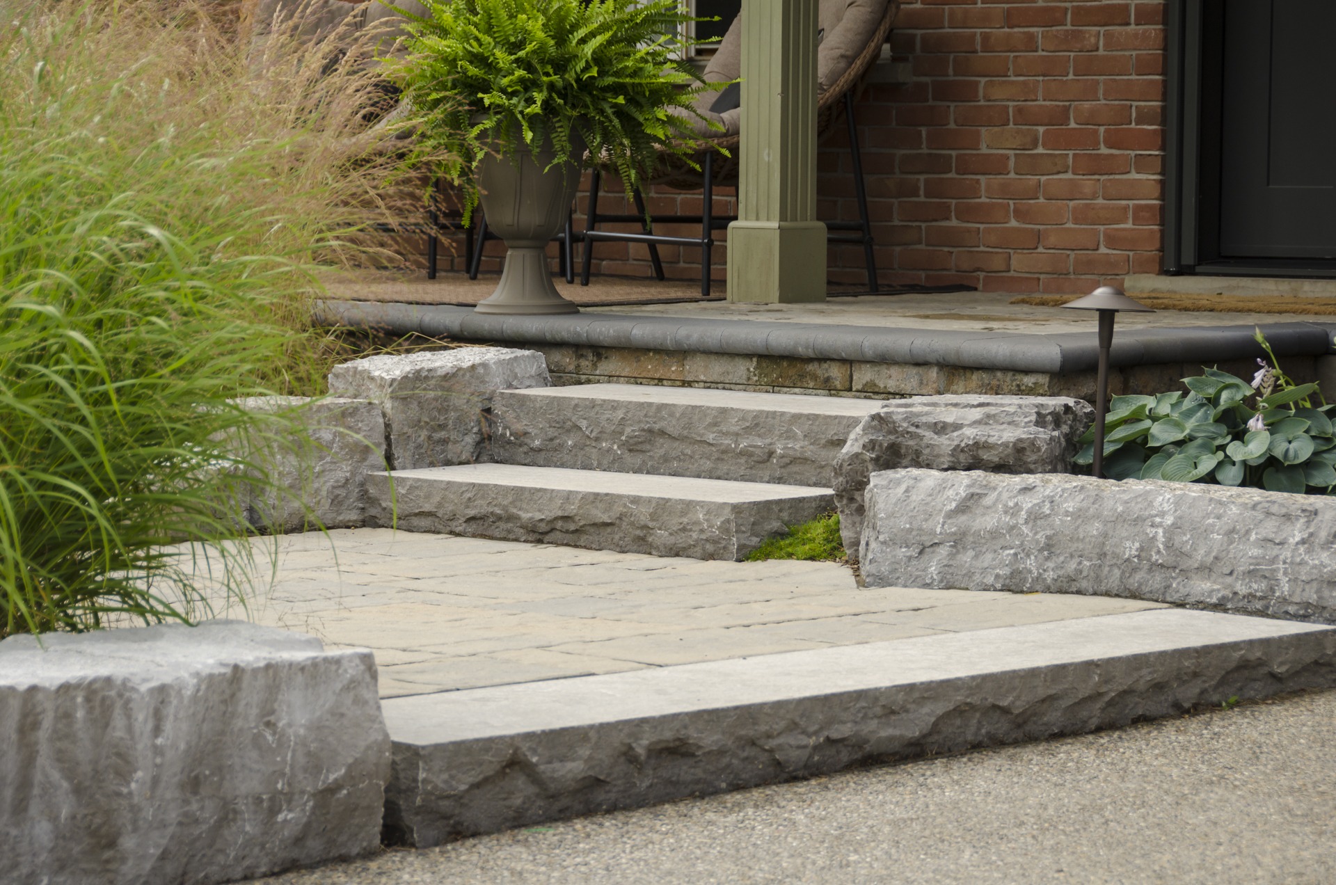 A stone stairway leading to a house bordered by green grass and lush plants, with a brick wall and fern-filled urn nearby.