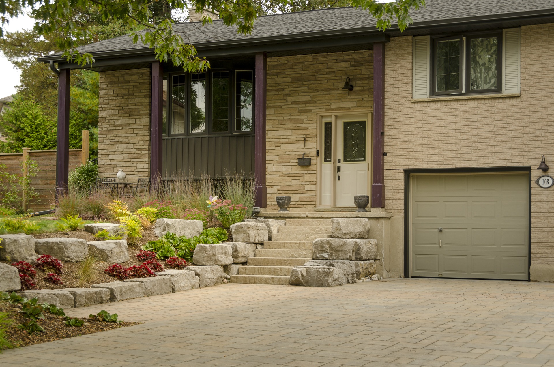 A stone facade house with a beige door, landscaped steps, and a single-car garage. Greenery and flowering plants adorn the front yard.