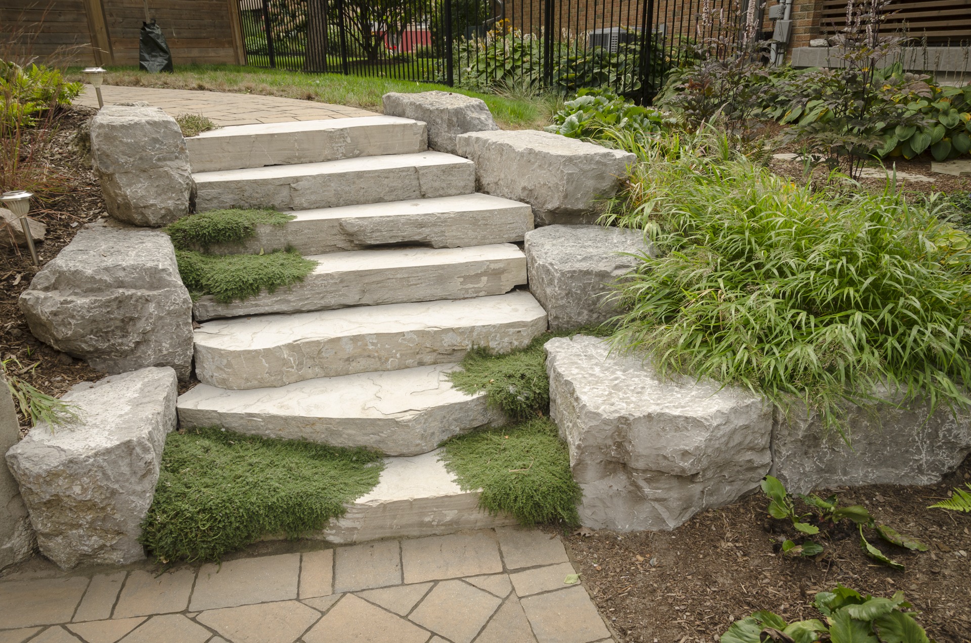 The image shows a beautifully landscaped garden with natural stone stairs featuring green ground cover plants between the steps, surrounded by lush vegetation.