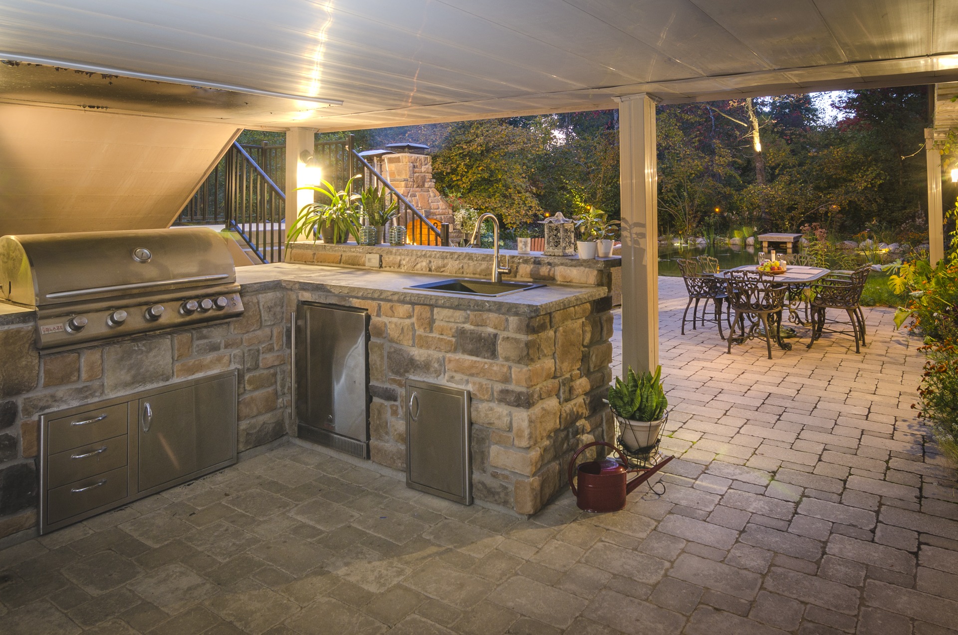 An outdoor patio with a stone-built grill station, under a covered area, leading to a garden with a dining set and lush greenery at dusk.