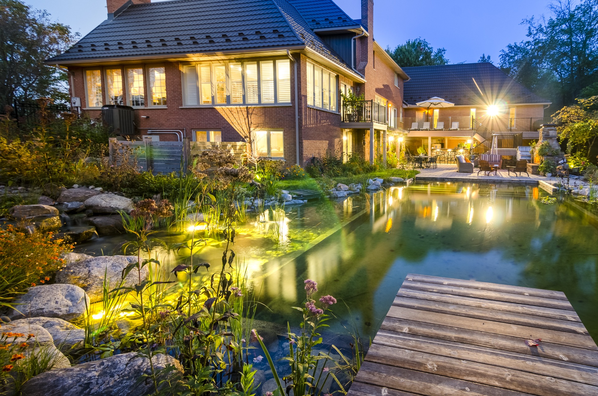 This image shows a twilight scene of a luxurious backyard with a natural pond, illuminated garden, wooden deck, and a cozy patio area.