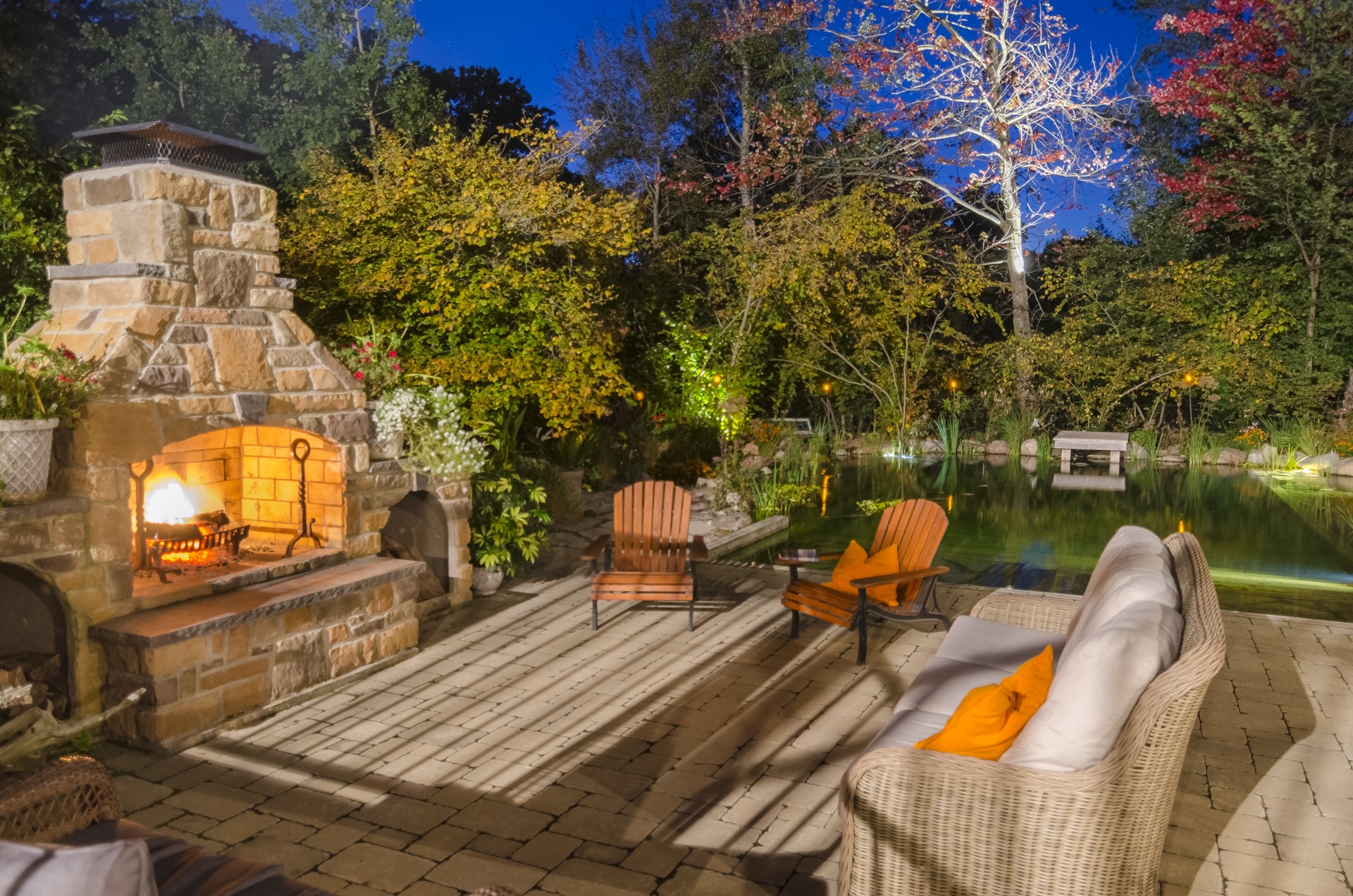 An inviting outdoor space at twilight with a lit stone fireplace, wooden chairs, comfy sofa, vibrant trees, and a peaceful pond under a clear sky.