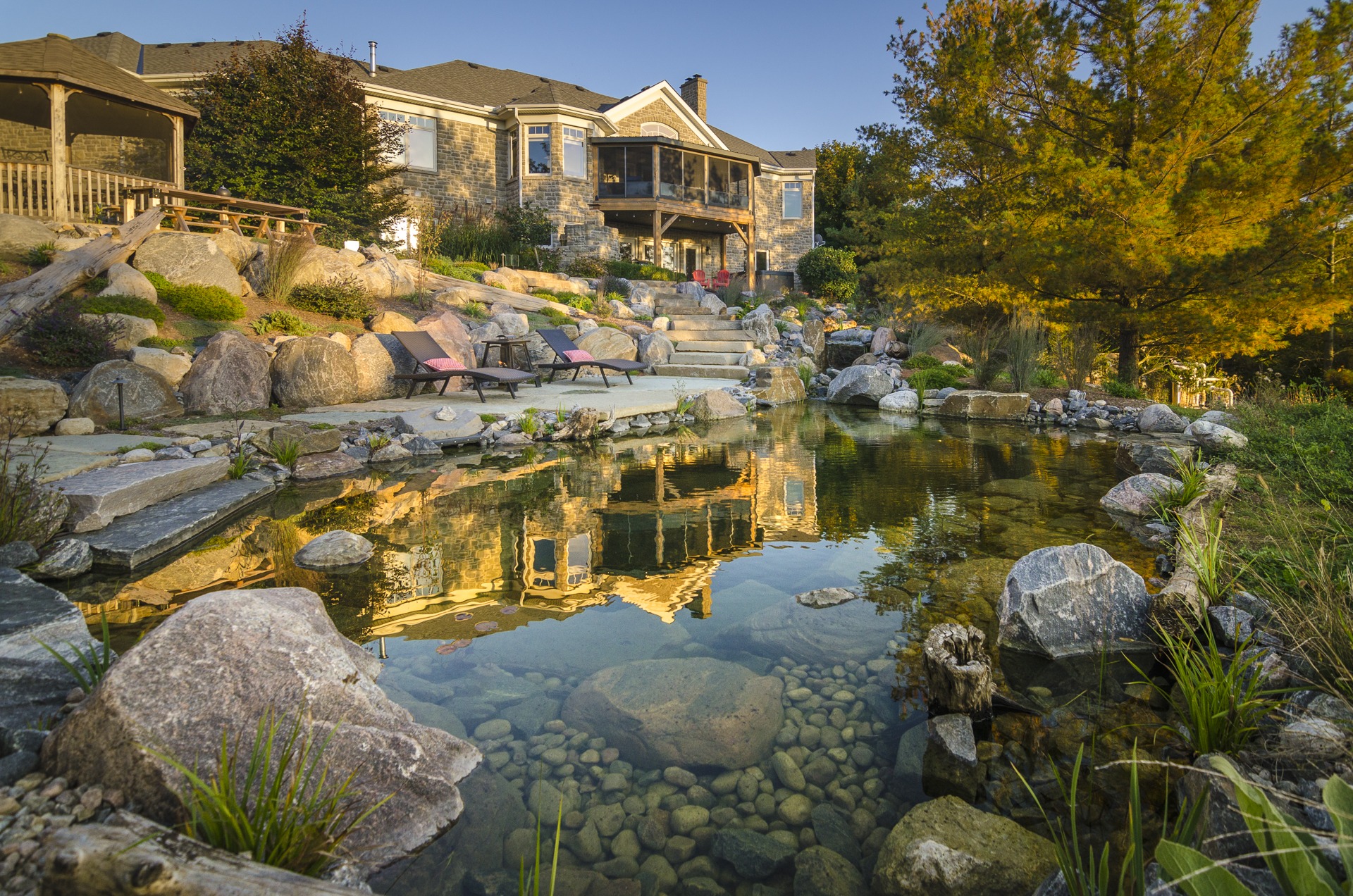 This image shows a tranquil backyard pond reflecting a stone house surrounded by lush vegetation, rocks, lounge chairs, and a clear evening sky.