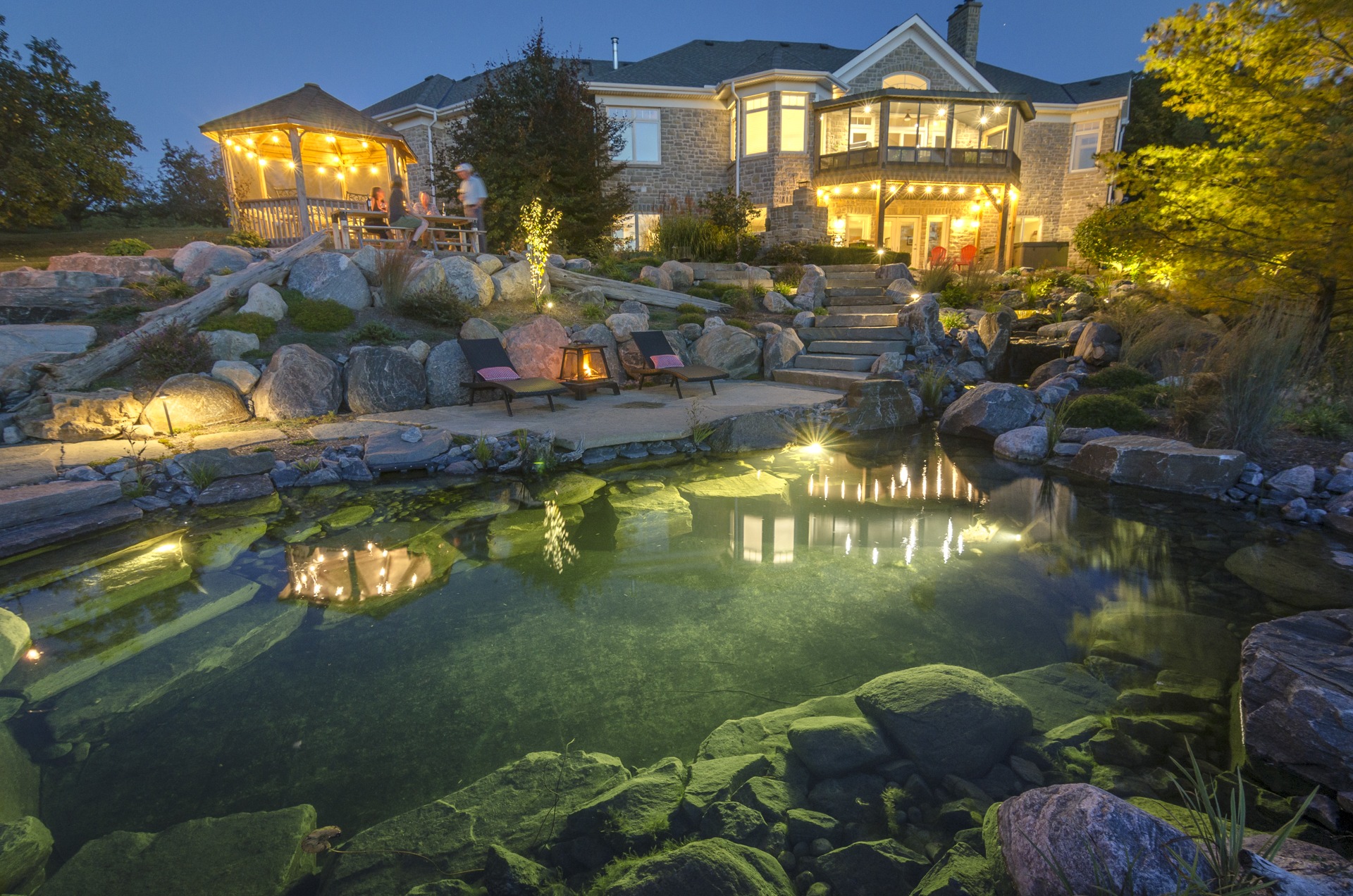 A luxurious house at dusk with warm lighting, featuring a landscaped garden, stone steps, a natural pond, and people enjoying an outdoor gazebo.