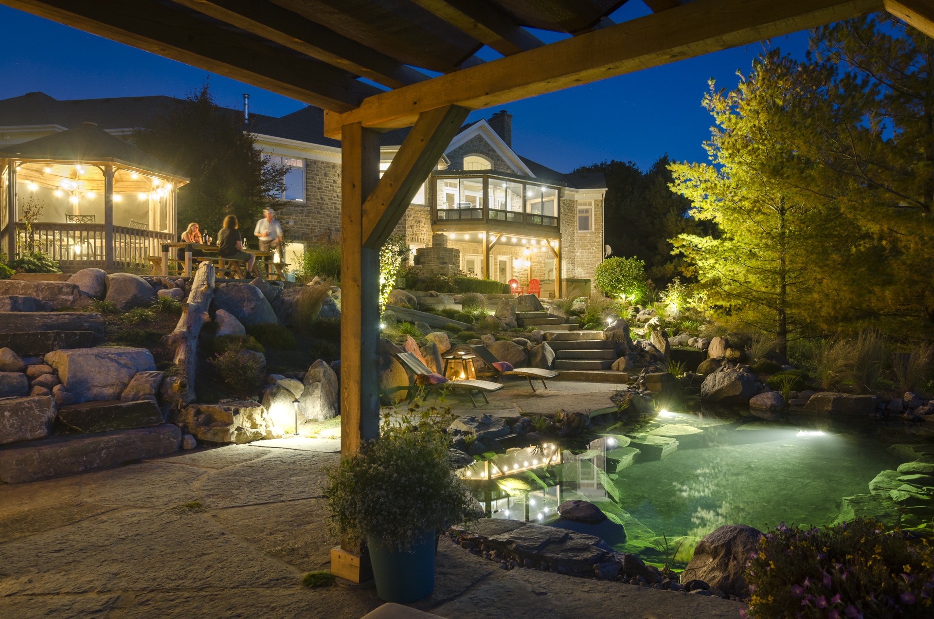 This image shows an illuminated backyard at dusk with a pond, stone landscaping, and a house. People gather on a deck with warm lighting.