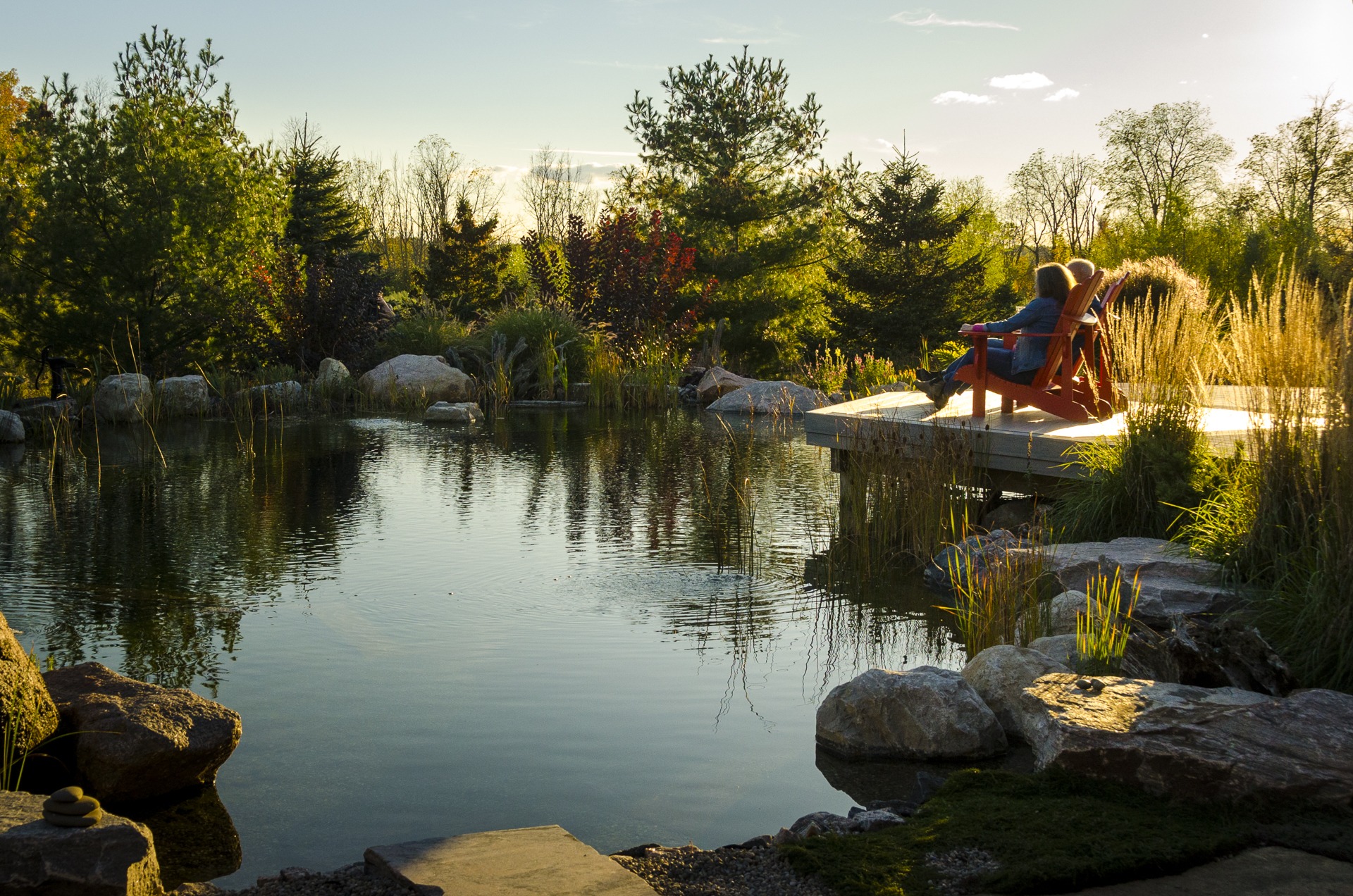 A serene pond surrounded by rocks and greenery, with a person relaxing on a red Adirondack chair on a wooden dock during sunset.