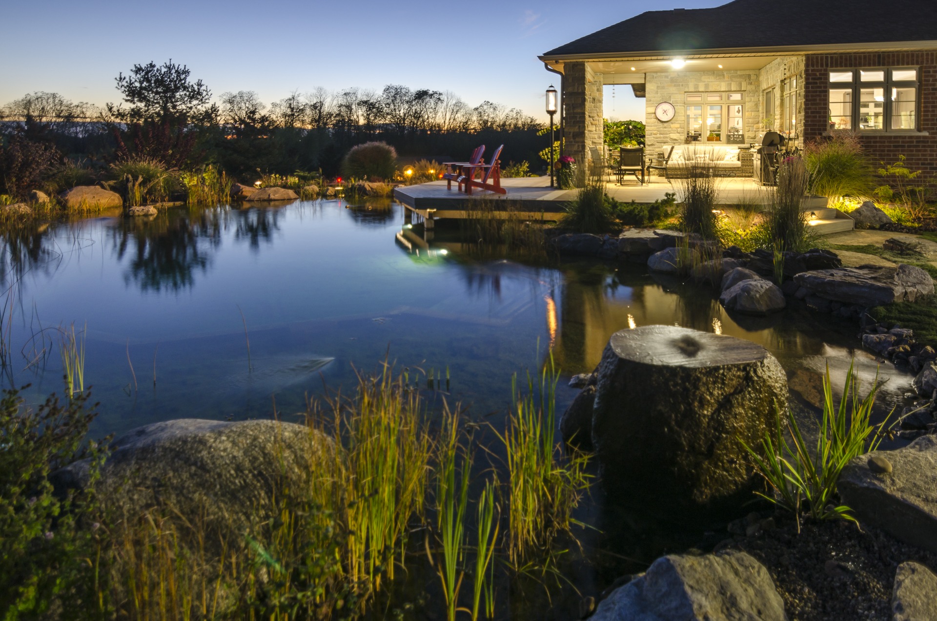 A serene twilight scene featuring a calm pond, reeds, rocks, a cozy brick house with lit windows, an inviting porch with furniture, and Adirondack chairs.