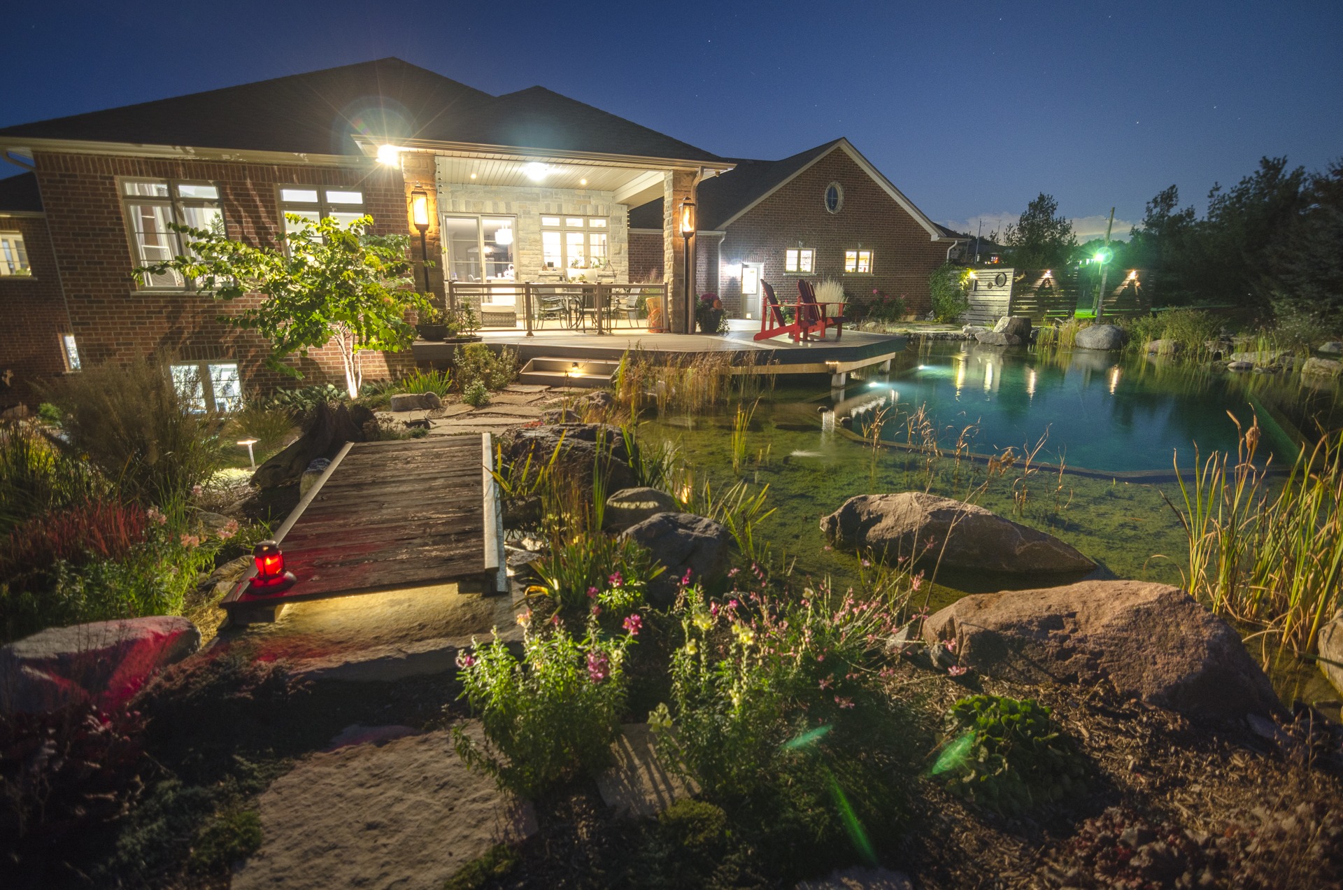 Nighttime view of a brick house with a lit deck overlooking a naturalistic garden pond. A wooden bridge, rocks, and plants enhance the serene setting.