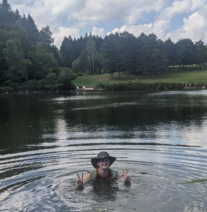 A person is submerged in water up to their shoulders, smiling, wearing a hat, and making peace signs, surrounded by a natural landscape with trees.