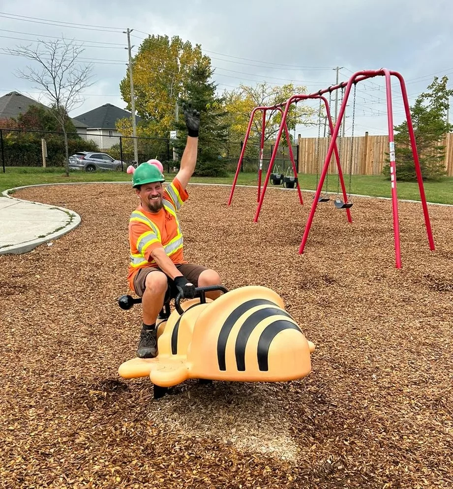 A person in a reflective vest and a colorful helmet happily raises their hand while sitting on a playground spring rider shaped like a bee. Swing set in the background.