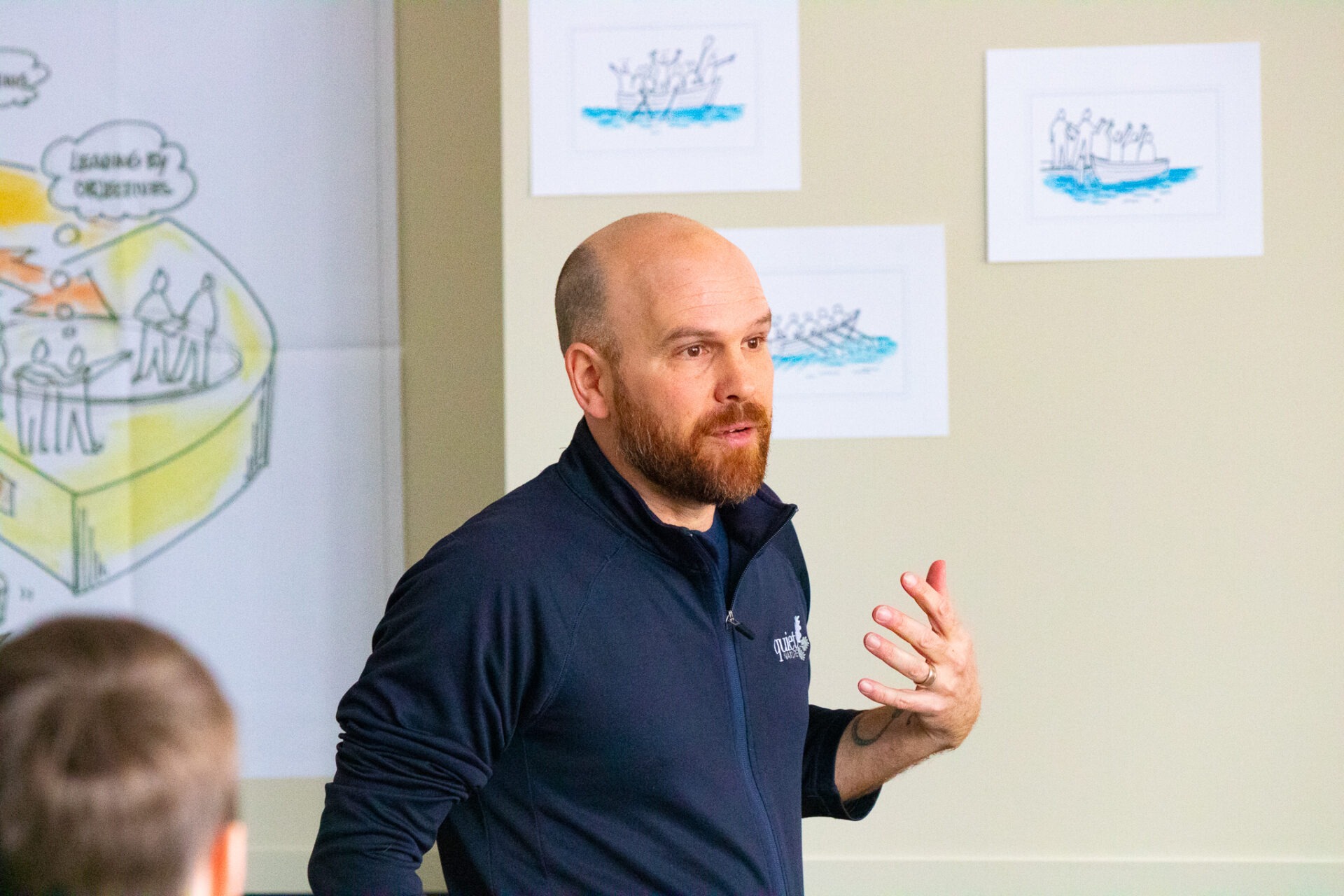 A person is actively speaking and gesturing with hand raised. Drawings of boats and teamwork diagrams are on the wall behind them. They wear a dark shirt.