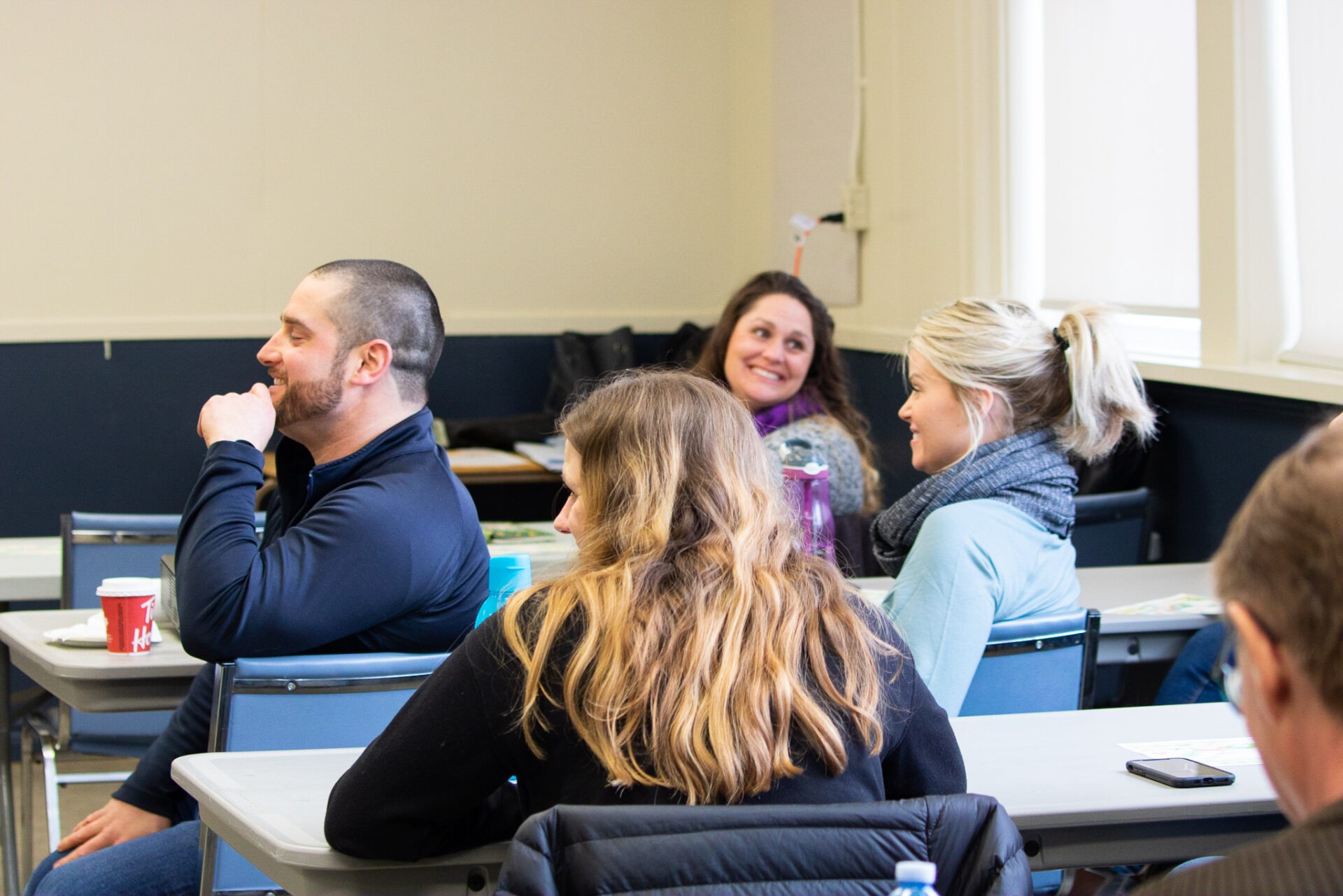 Four adults are sitting in a classroom setting, chatting and smiling, with coffee cups and water bottles on their desks.