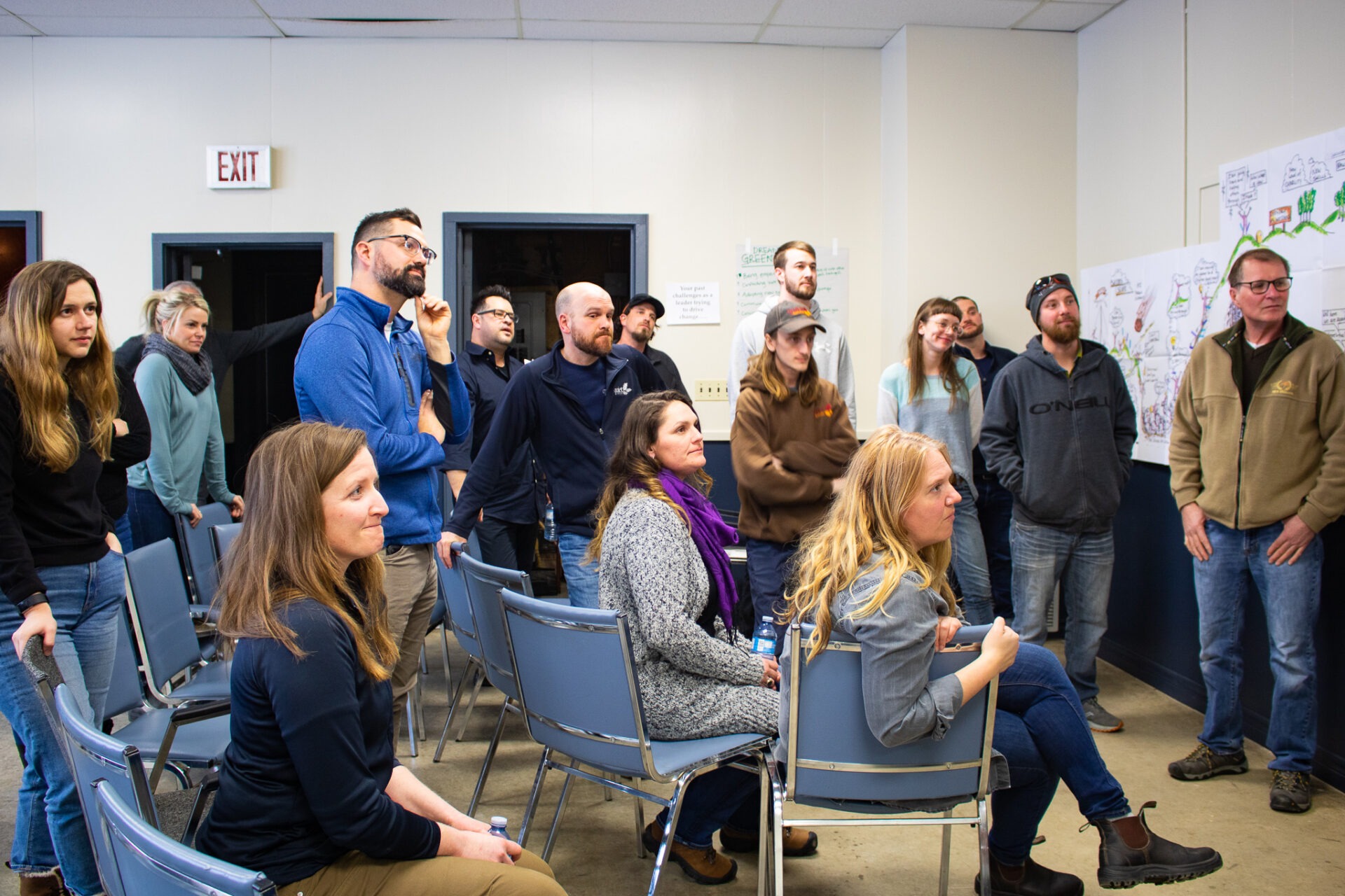 A group of people is attentively standing and sitting in a room with chairs, looking towards something off-camera, possibly during an event or meeting.