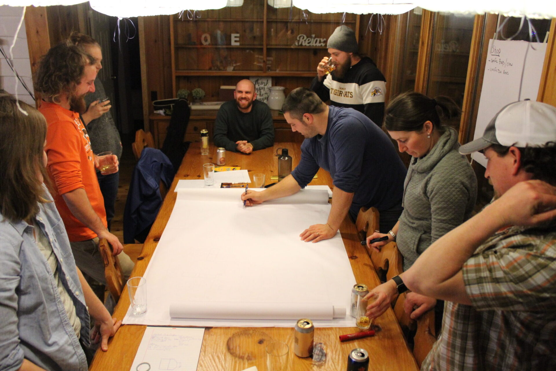 A group of people gather around a table, some standing, some sitting. One person is drawing on large paper. The room looks casual and cozy.