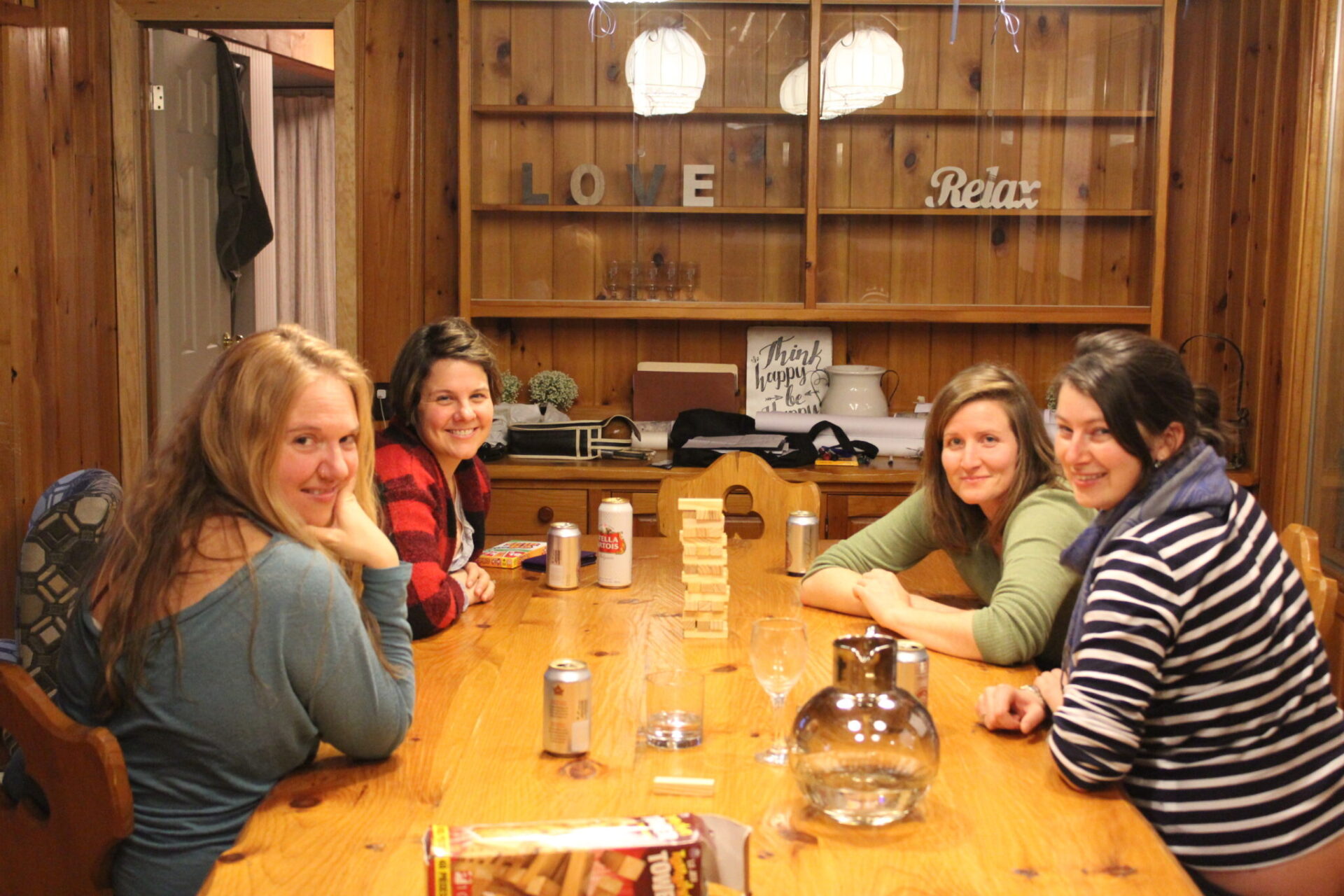 Four people sit around a wooden table with a Jenga game set up, smiling at the camera, in a cozy room with 