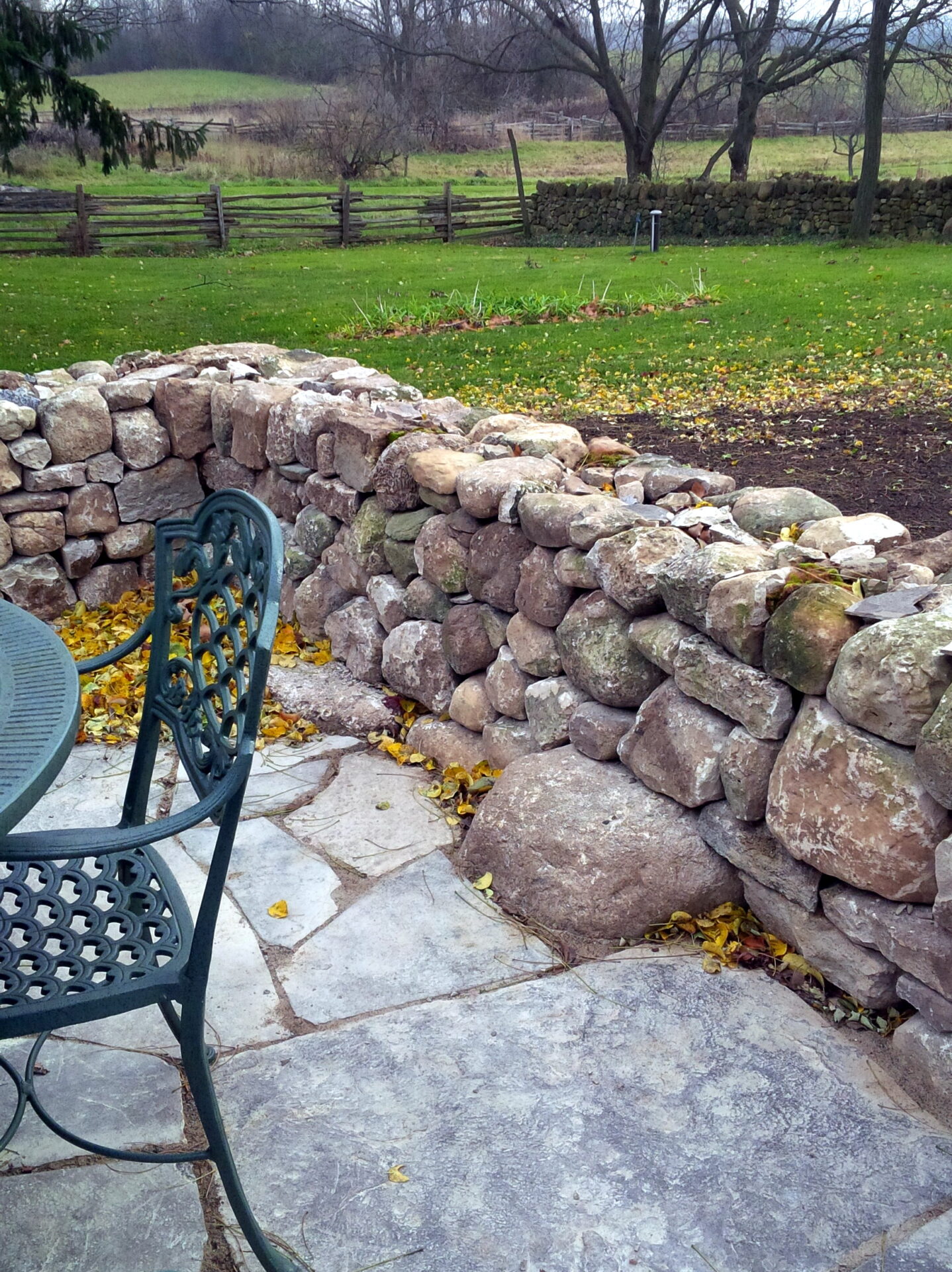 A rustic patio with a stone retaining wall and a metal chair overlooks a serene lawn bordered by a wooden fence and autumn foliage.