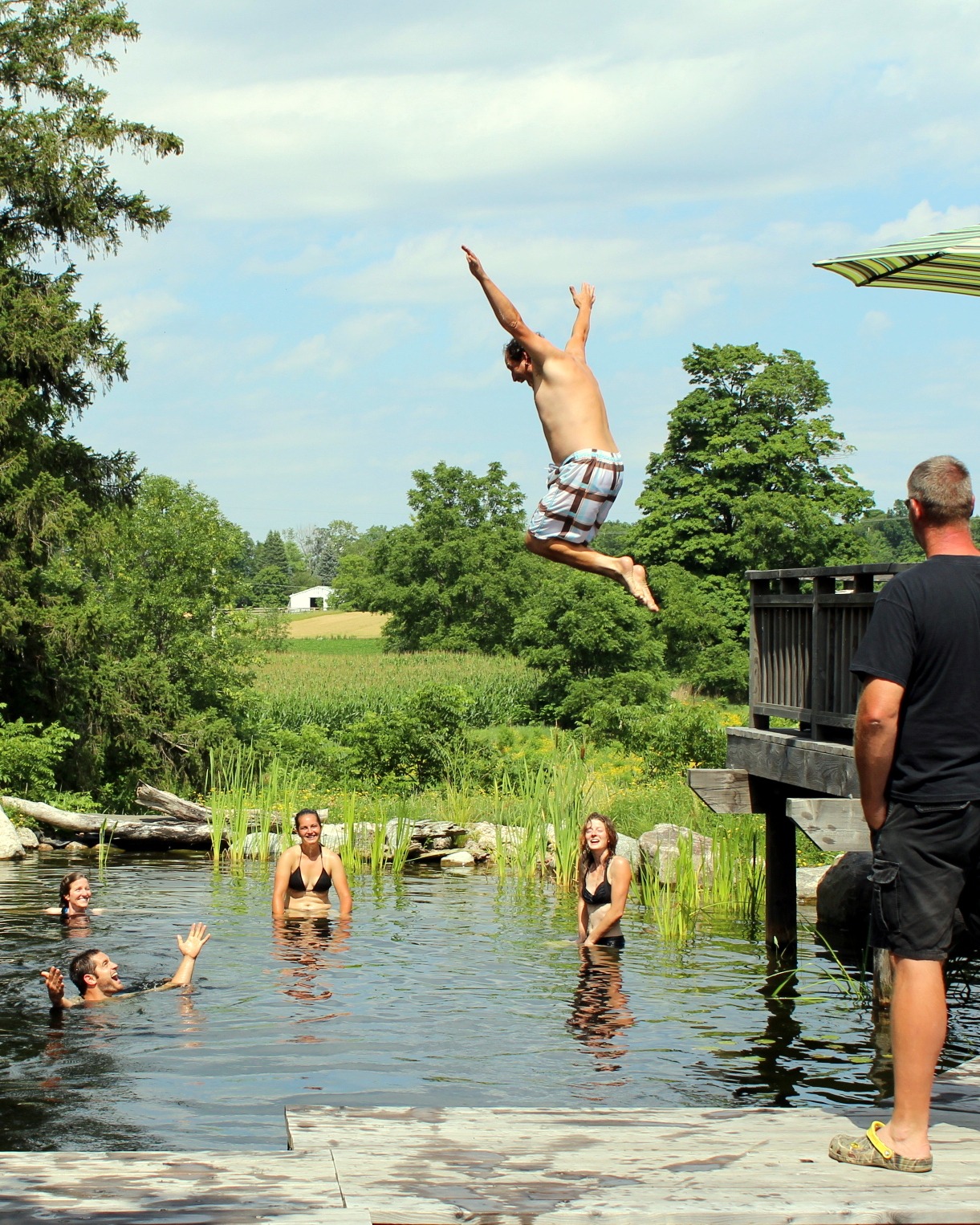 A person is mid-air, backflipping into a pond, while others watch and wait in the water. Trees, sky, and a rural backdrop round out the scene.