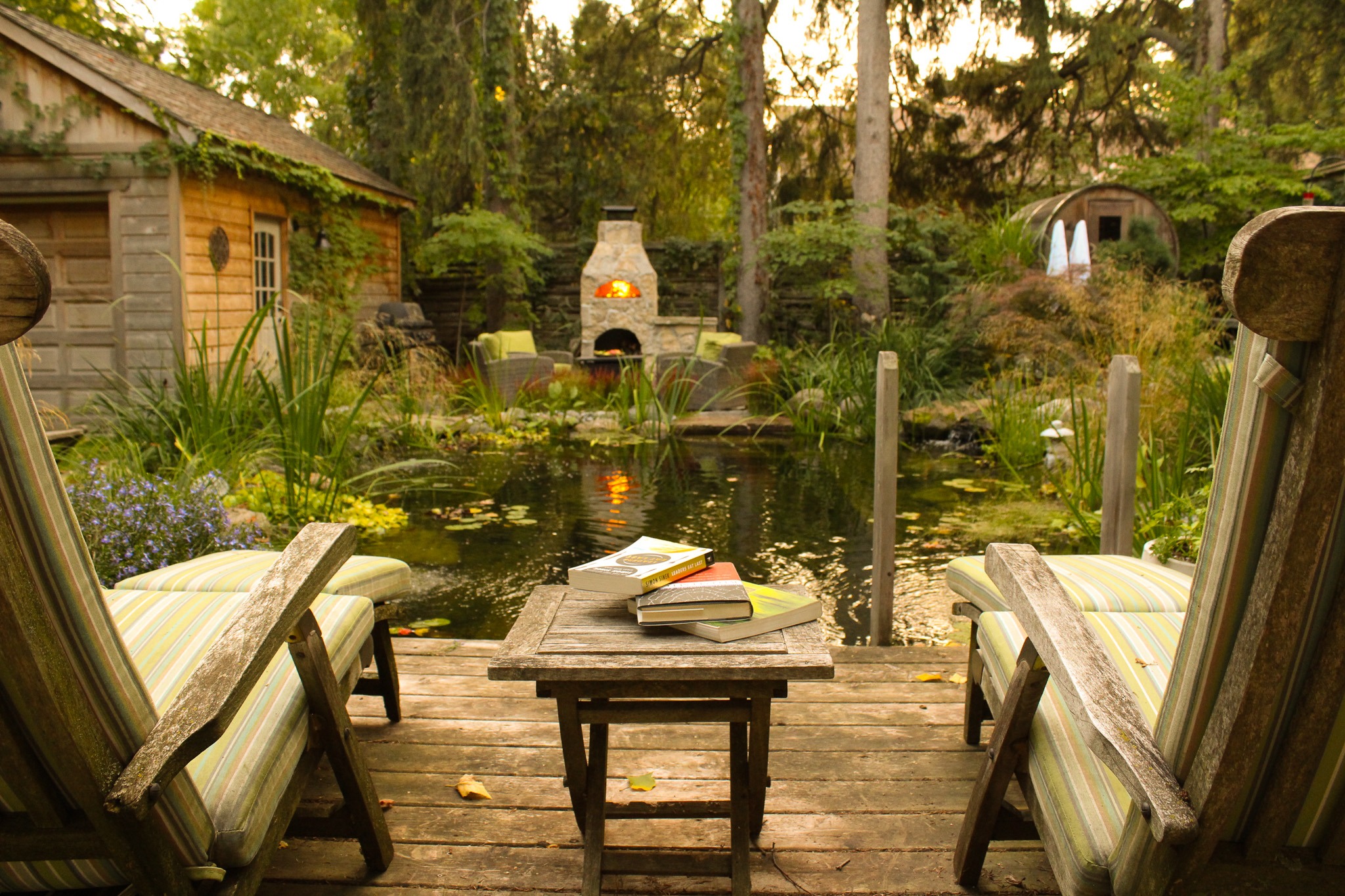 A cozy outdoor setting featuring two Adirondack chairs facing a serene pond, with a small table holding books, and a lit fireplace in the background.