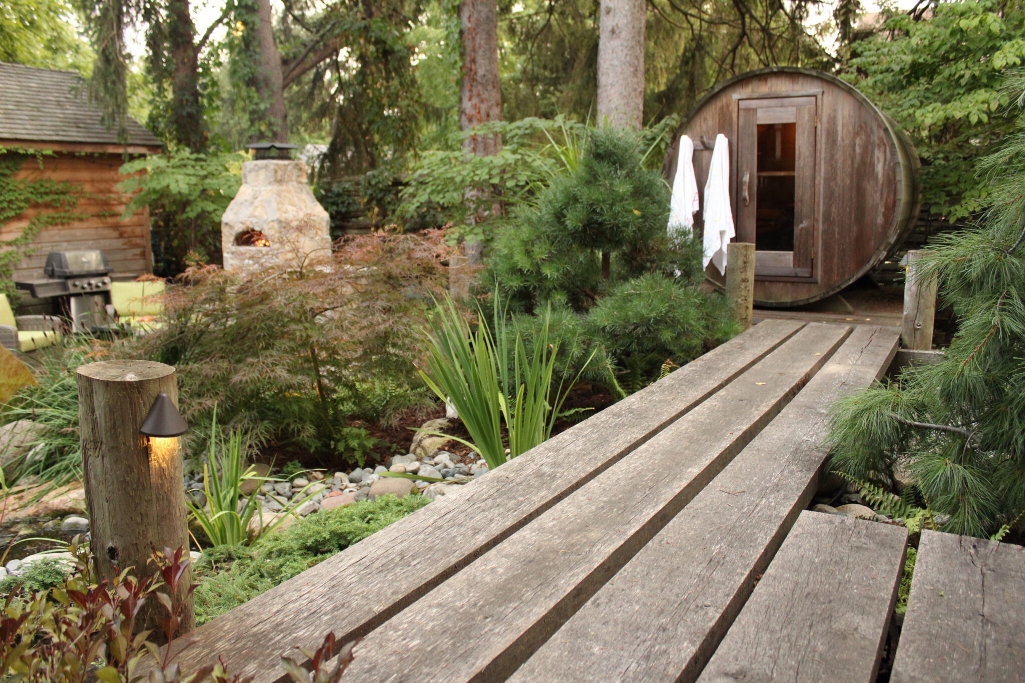 An outdoor space featuring a wooden deck, landscaped garden, an open outdoor fireplace, BBQ grill, and a barrel-shaped sauna in a serene woodland setting.