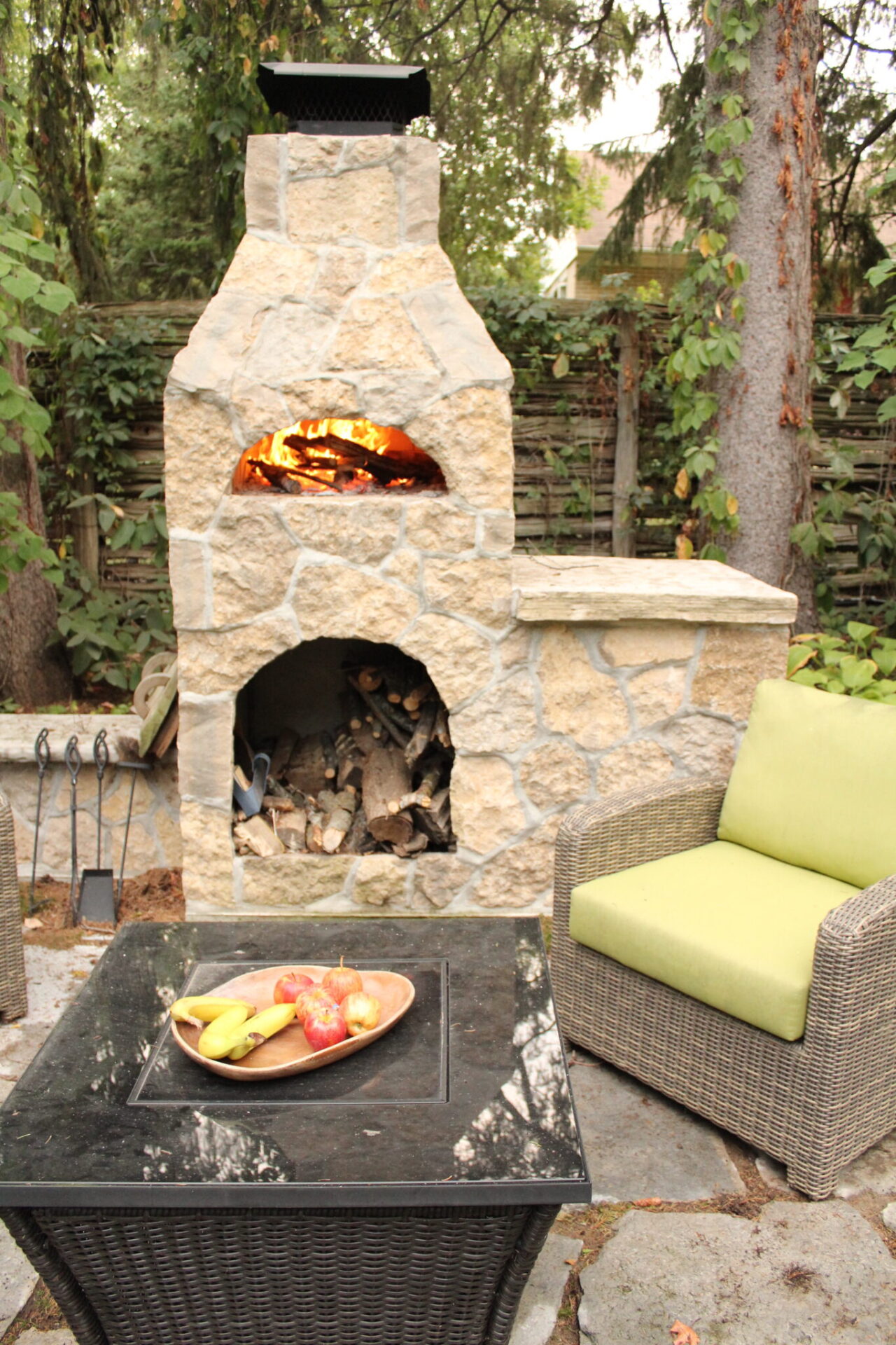 An outdoor stone fireplace is ablaze with a firewood storage underneath. A wicker chair with a green cushion and table with fruit rest nearby.